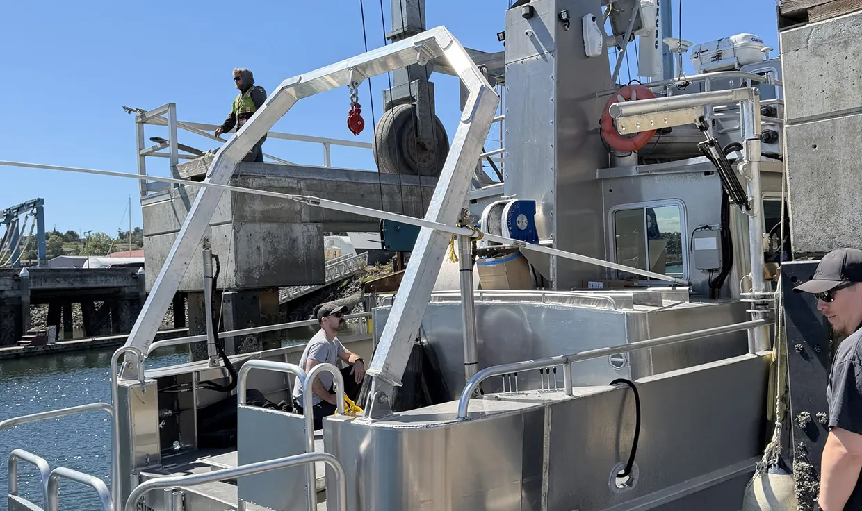Two men work on a small silver research boat docked by an industrial pier on a sunny day, with one man sitting inside and another standing on the dock above.