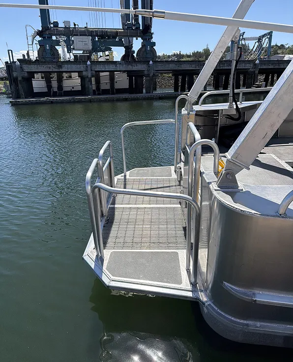 Metal boarding platform with railings extending from the stern of a boat over calm water, with industrial dock equipment and a clear blue sky in the background.