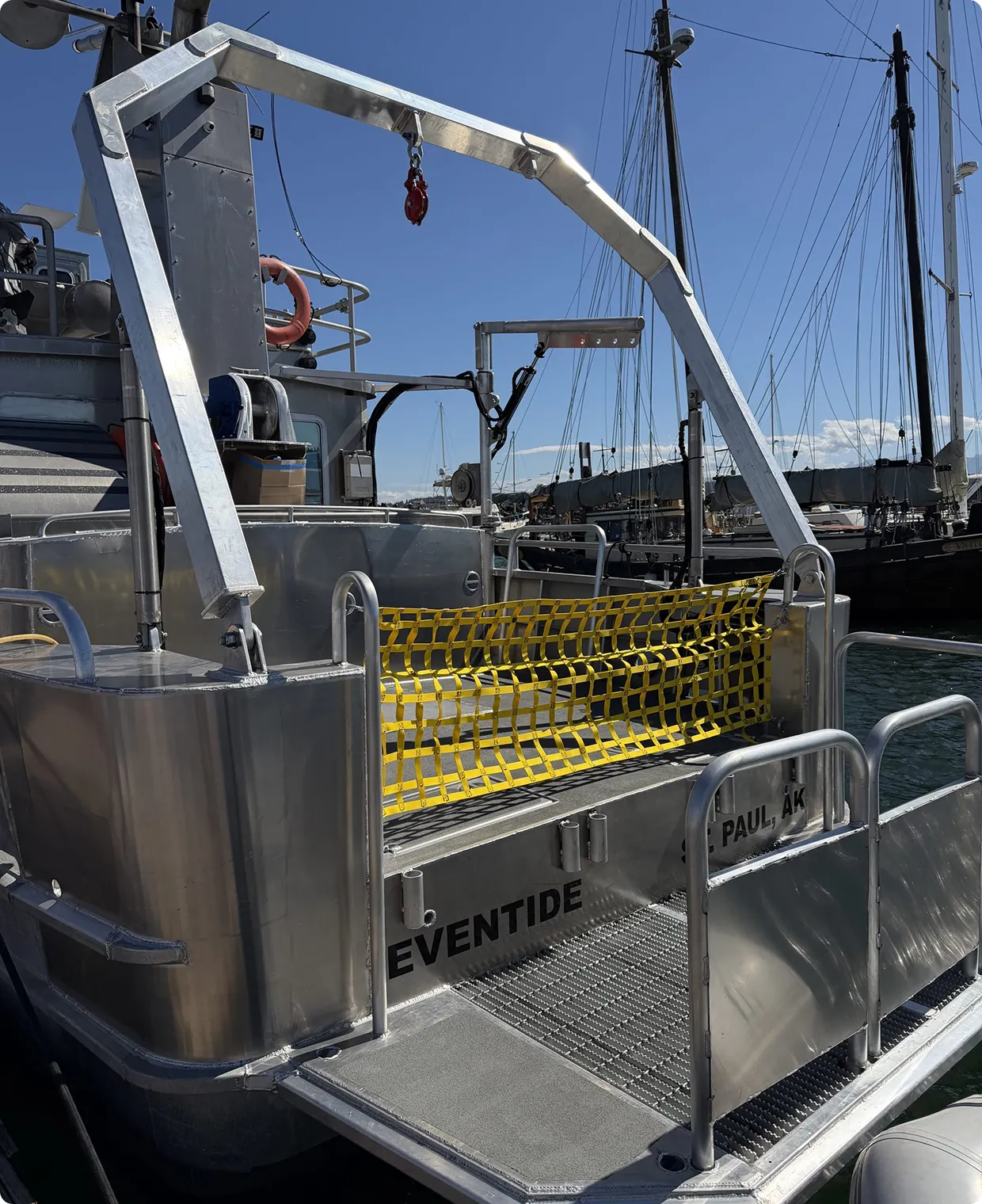 Rear deck of a silver metal boat named Eventide with a yellow safety net across the back opening, docked among other boats under a clear blue sky.