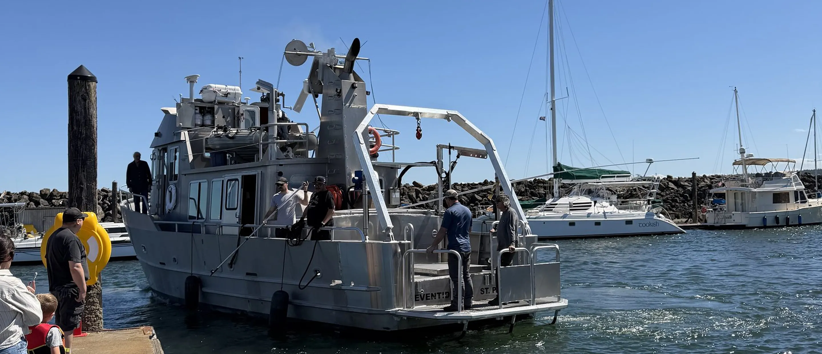 A silver metal boat docked at a harbor with five people visible on and near it, including two men waving. Nearby are other sailboats and a rocky breakwater under a clear blue sky.