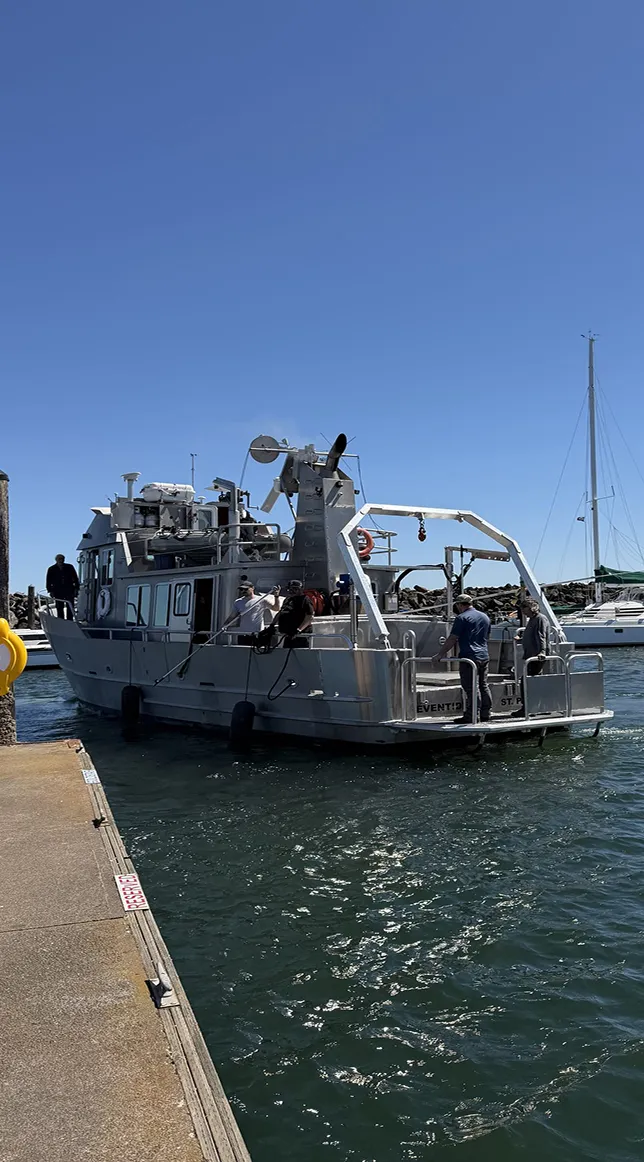 A metal research or work boat with four people aboard is docked at a pier in a harbor with calm water and clear blue sky.