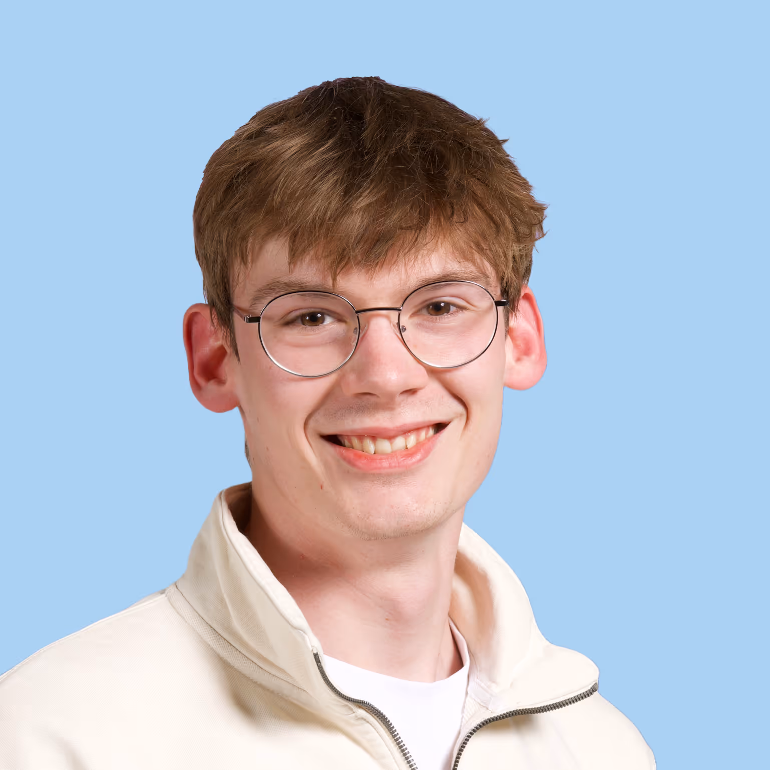 Young man with light brown hair, wearing round glasses and a cream zip-up jacket, smiling against a light blue background.