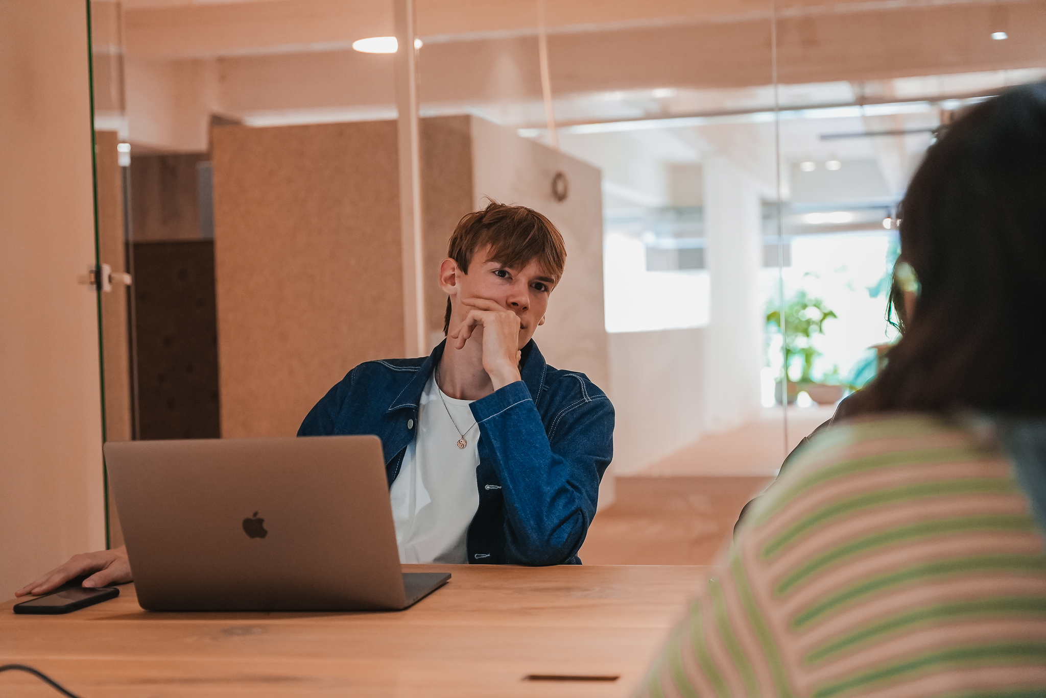 Young man in denim jacket sitting at a table with a laptop, resting his chin on his hand, looking thoughtfully at another person.