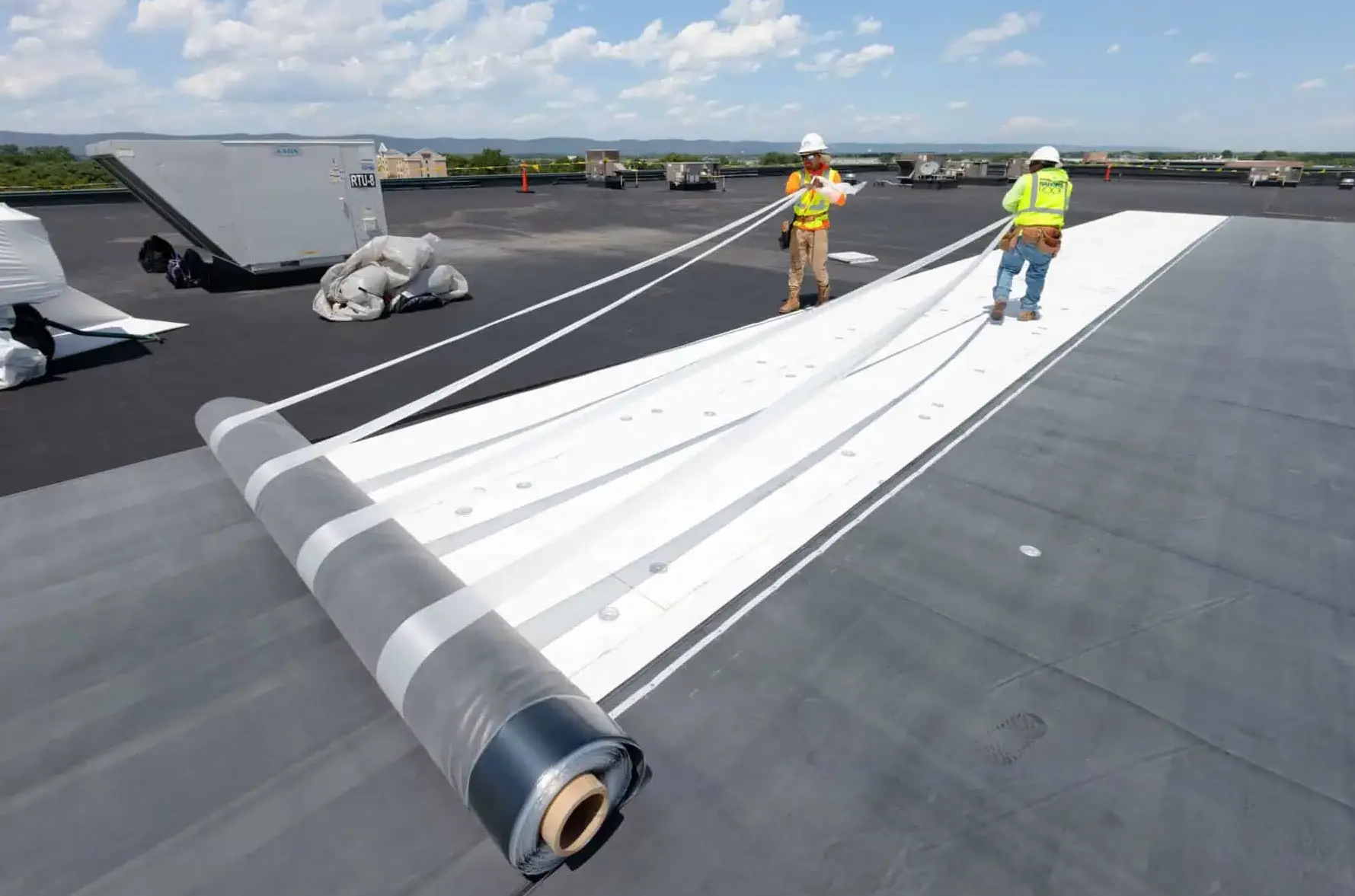 Two workers in safety gear unrolling a large white roofing membrane on a flat rooftop under a partly cloudy sky.