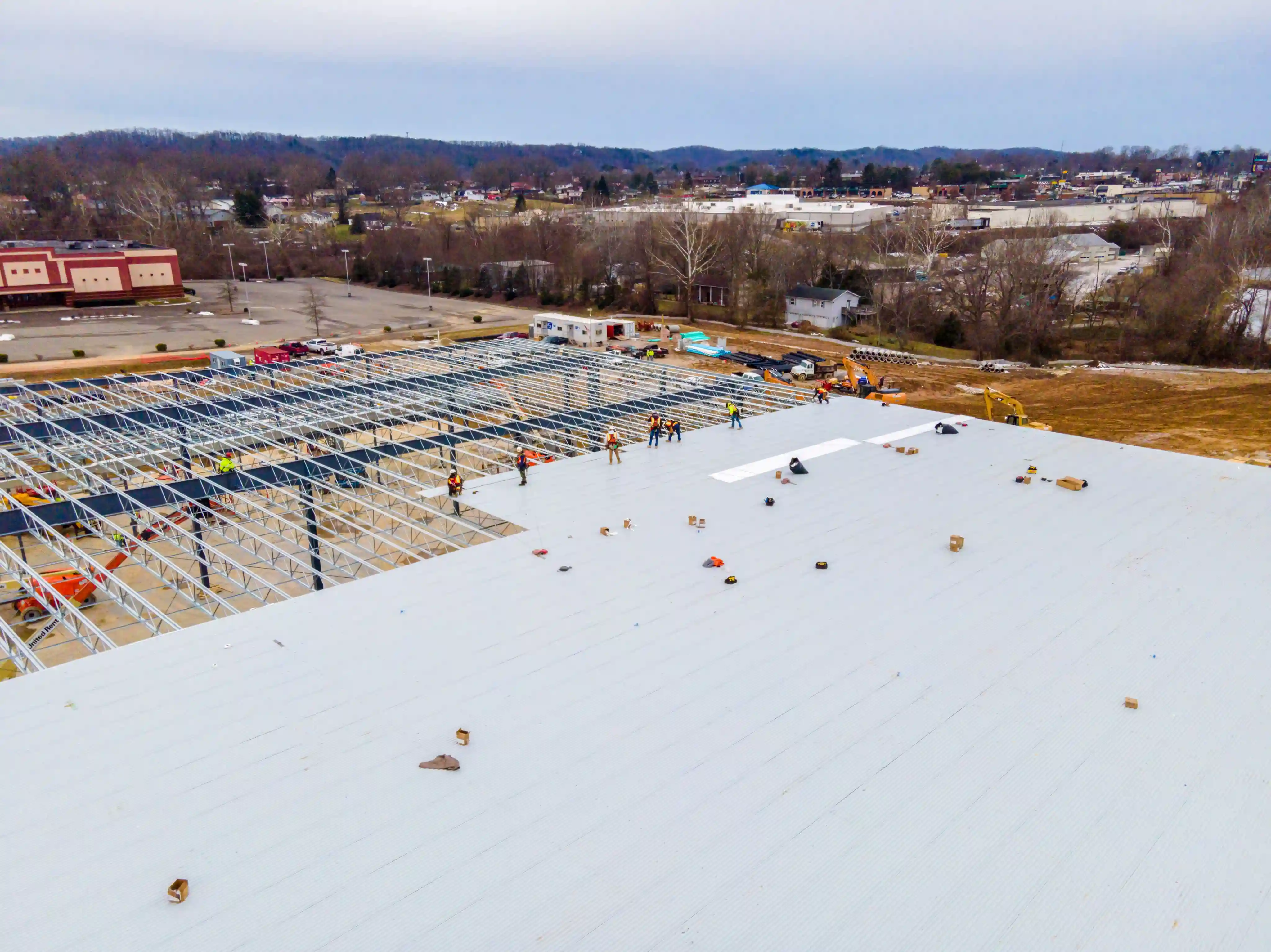 Aerial view of a construction site with workers installing a large white metal roof over a steel framework.