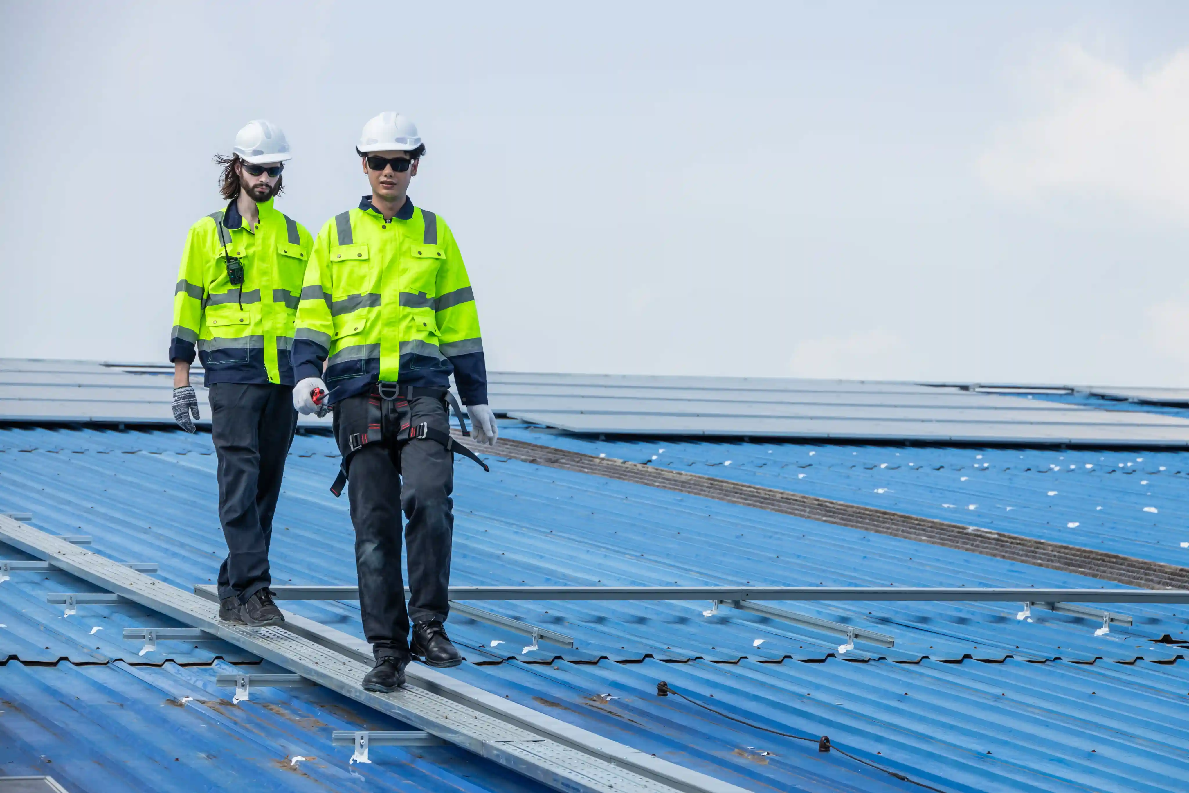 Two construction workers wearing white helmets and neon yellow safety jackets walking on a blue metal roof.