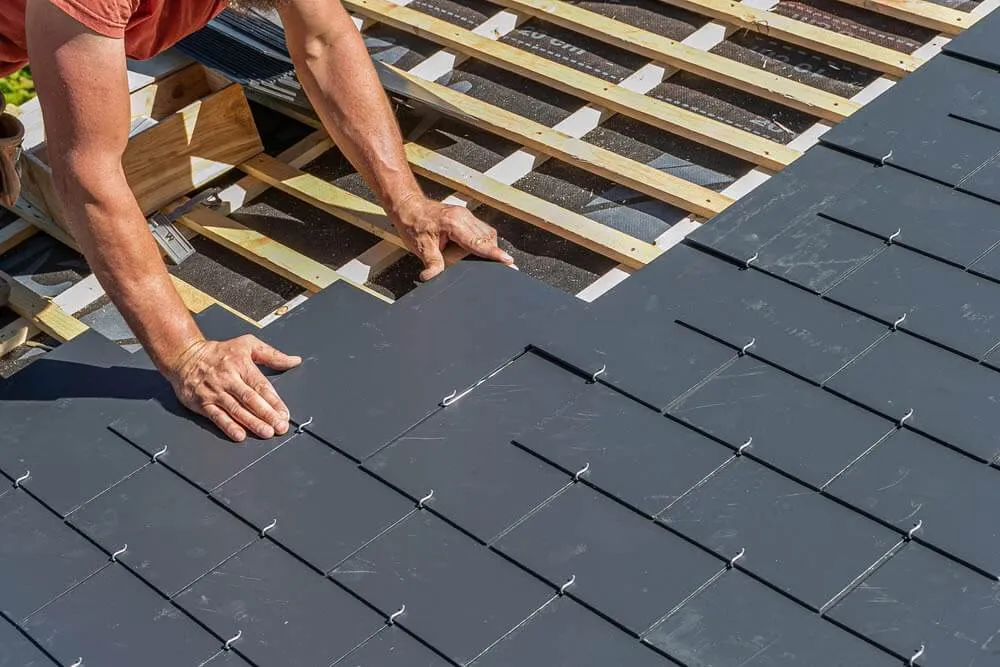 Construction worker installing overlapping dark gray roof tiles on wooden framework.