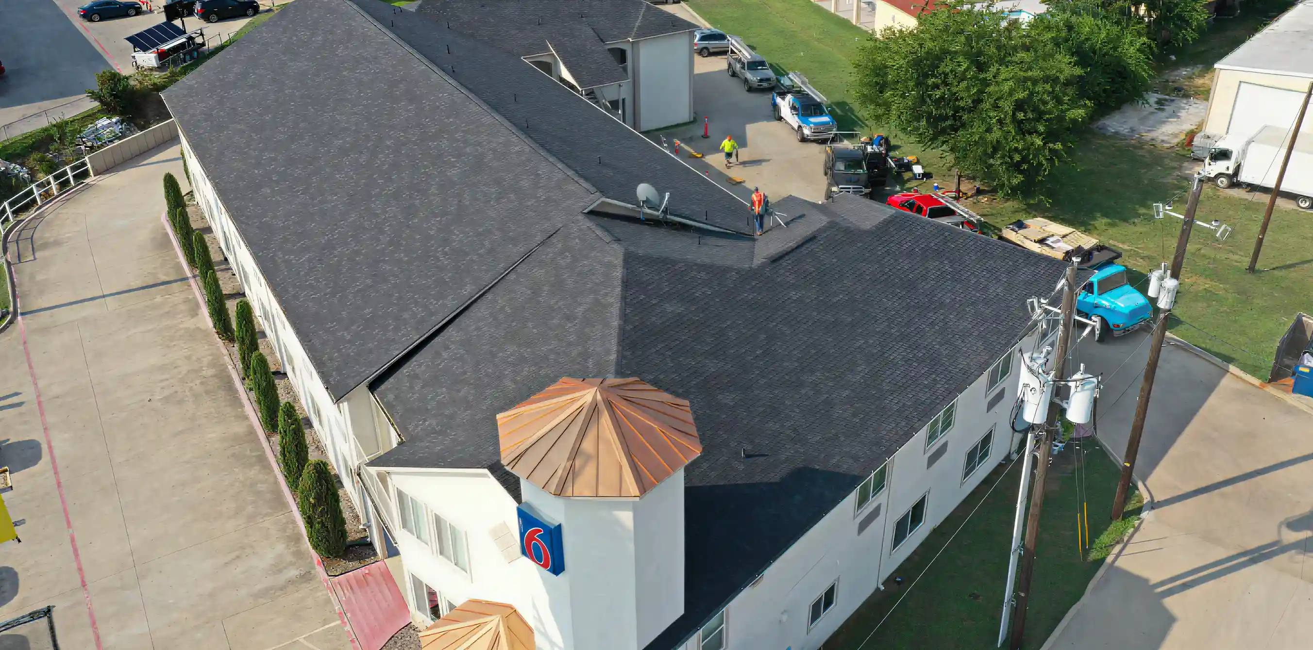 Aerial view of a two-story motel with a dark shingle roof, white walls, and a blue and red Motel 6 sign on a corner tower.