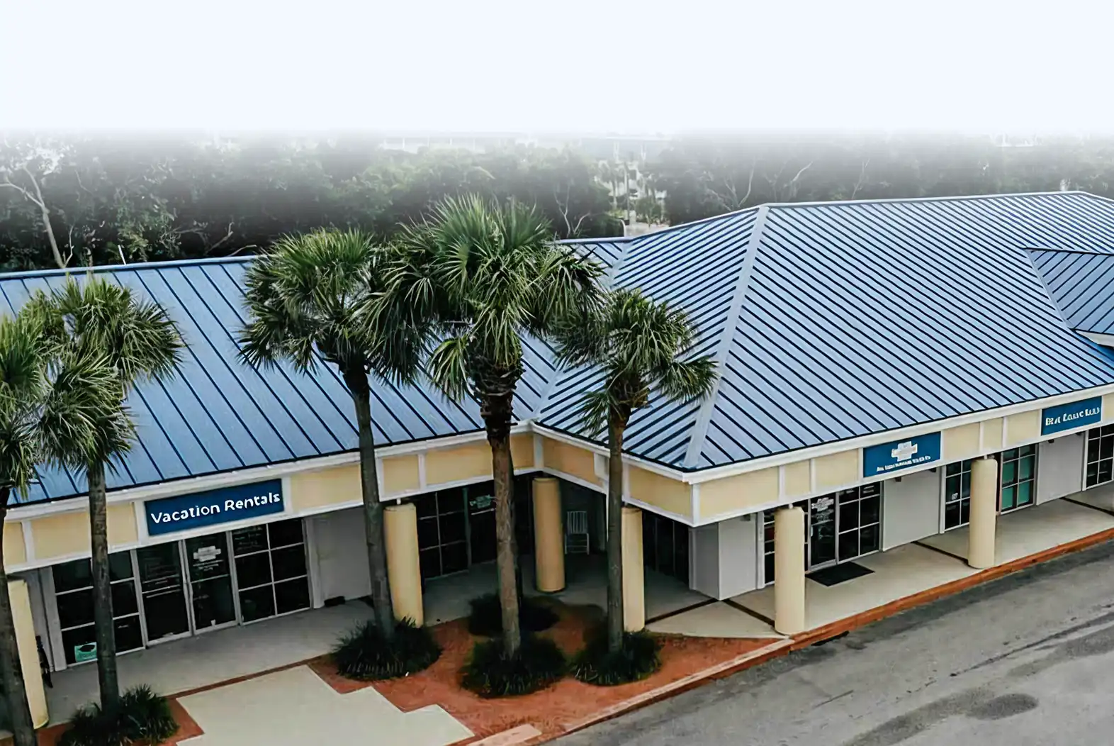 Small commercial building with blue metal roof and palm trees in front, housing businesses including a vacation rentals office.