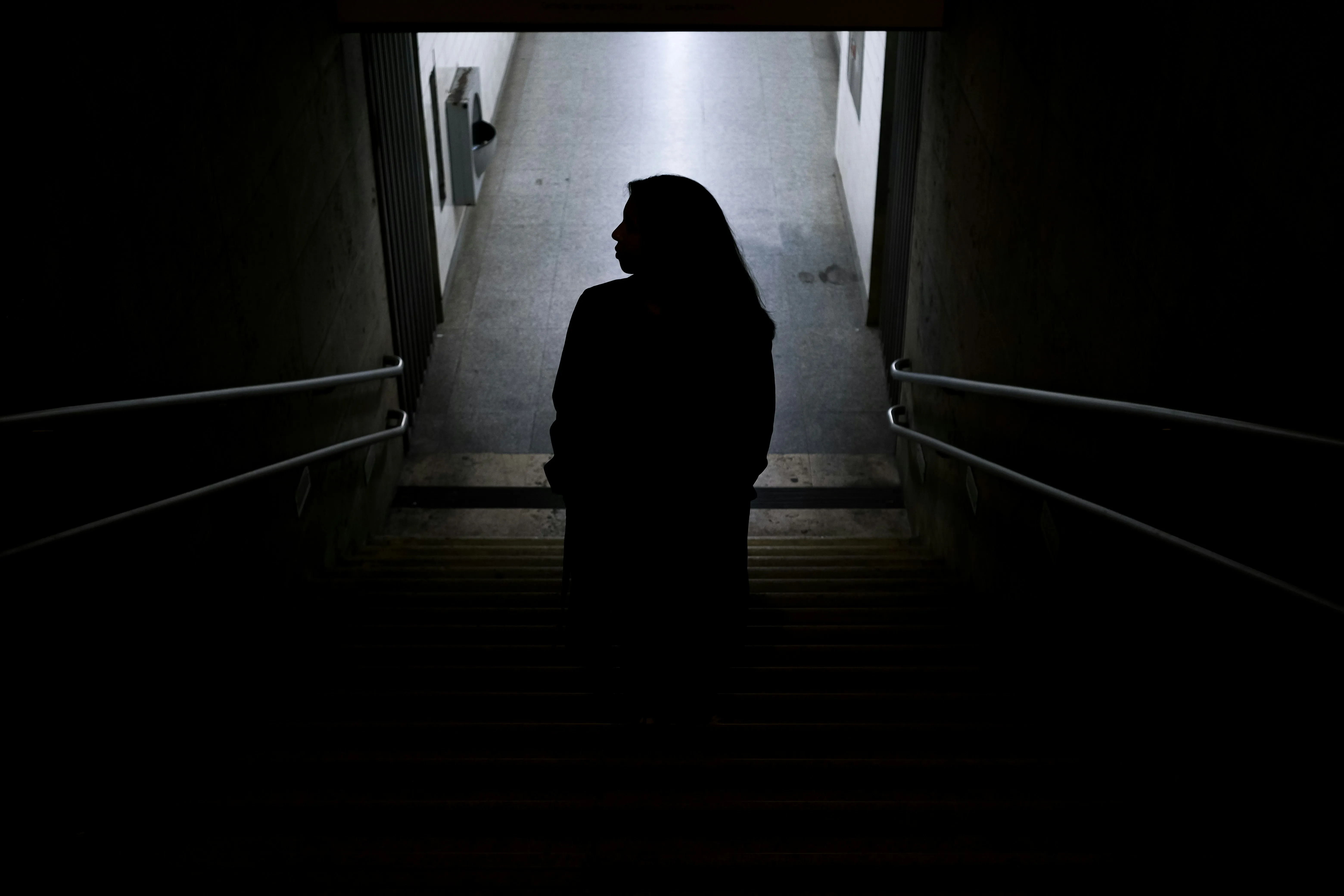 Silhuett av en person som står på en mörk trappa med händerna i fickorna och blickar åt sidan mot en ljus korridor.