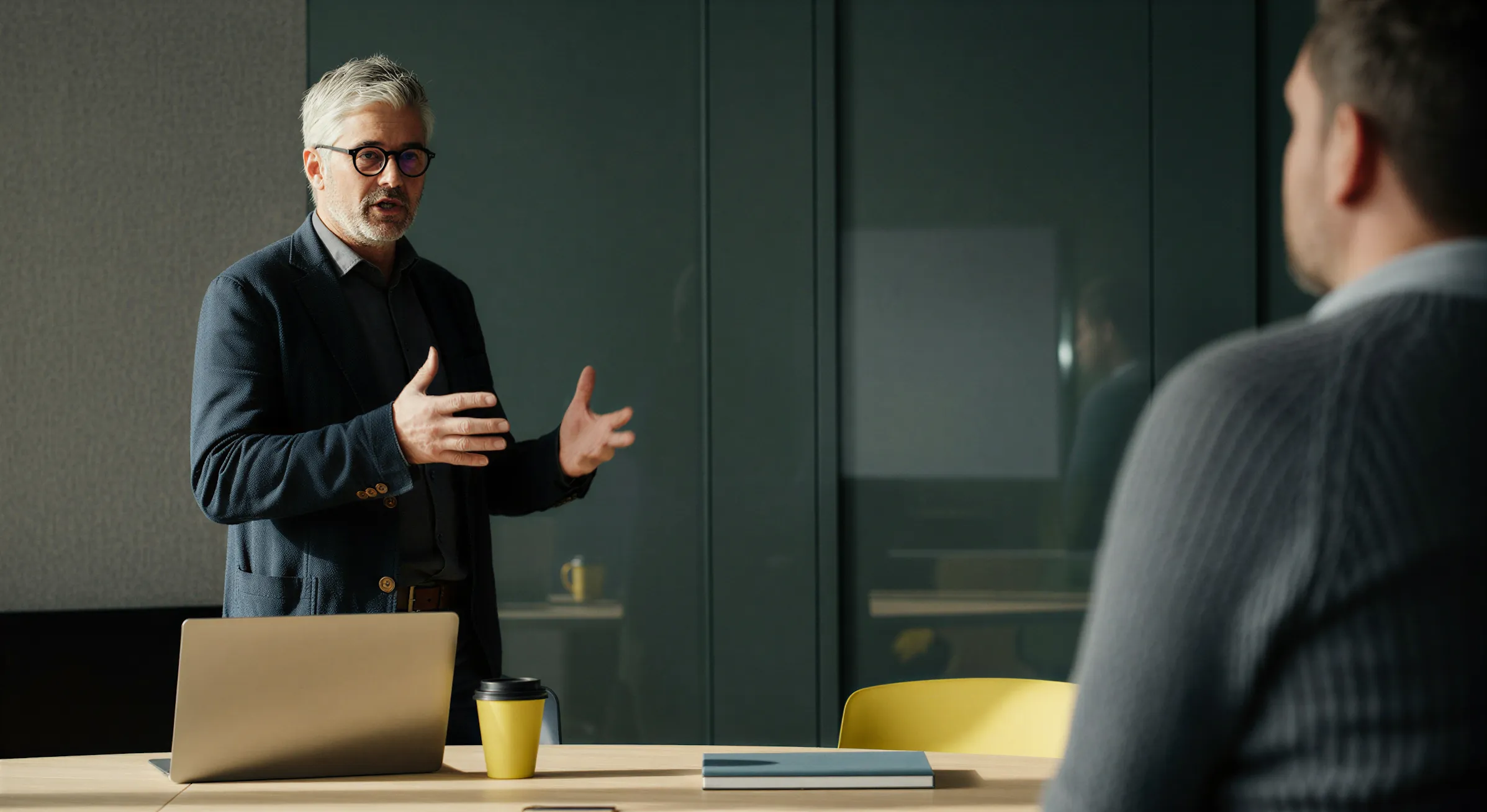 Man med grått hår och glasögon som gestikulerar vid ett bord med bärbar dator och kaffemugg medan han pratar med en annan person.