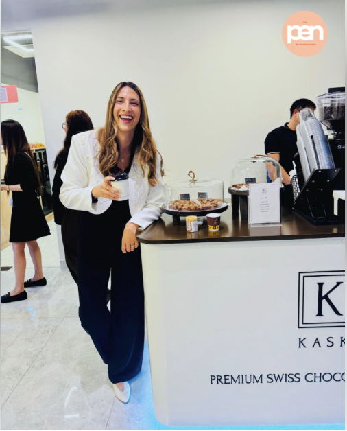 Smiling woman who is a business matchmaker in a white blazer holding a cup, leaning on a counter at a premium Swiss chocolate brownie popup stand inside a co-working community in Dubai with treats and a coffee machine.