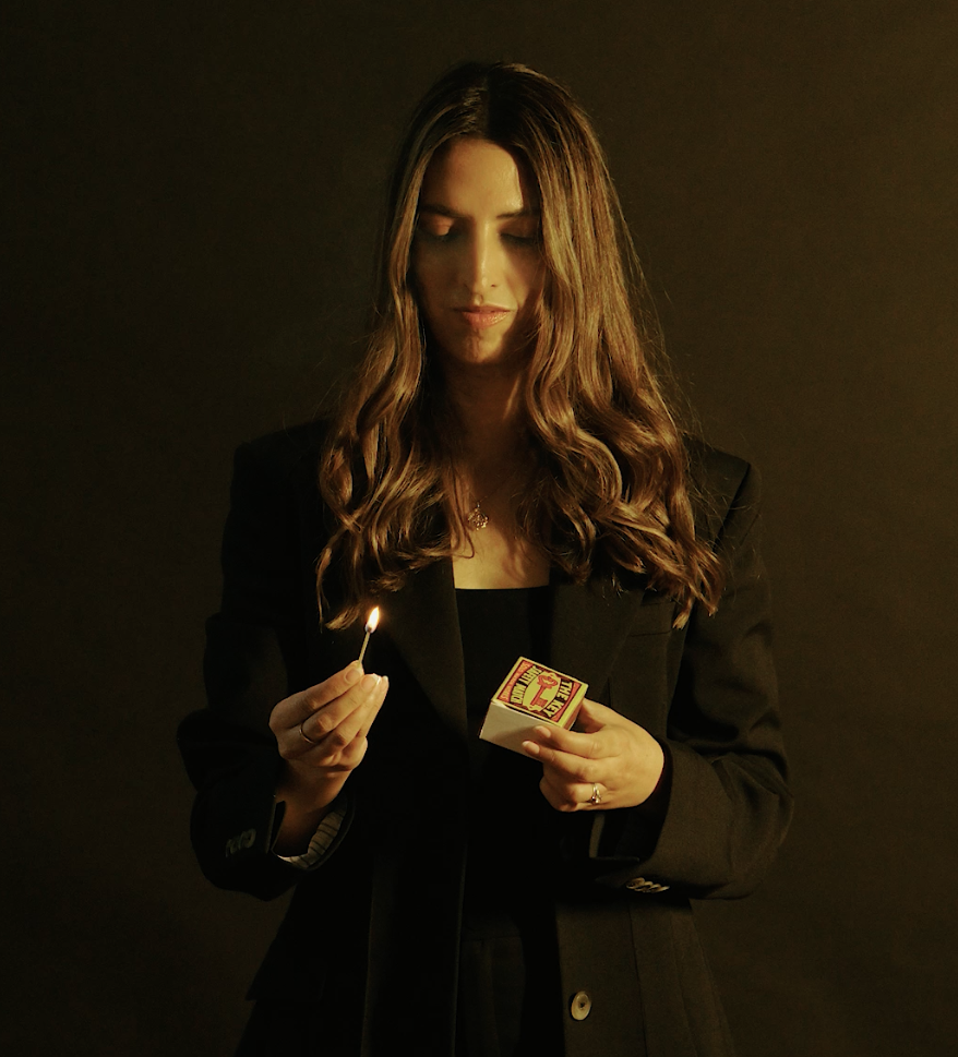 Woman who is a business matchmaker with long wavy hair wearing a black blazer holding a lit matchstick and a box of matches against a dark background.