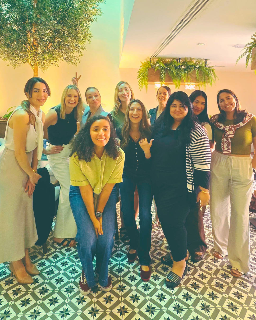 Group of ten smiling female founders posing indoors in a cafe in Dubai on a patterned tile floor with plants hanging and a tree in the background.