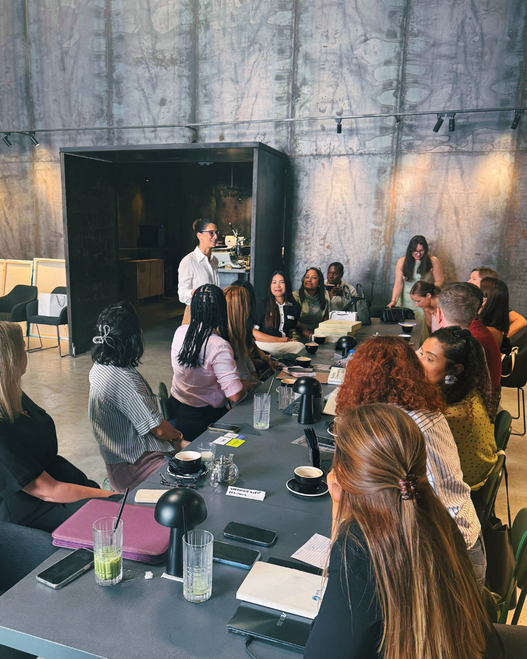 Group of diverse founders in Dubai gathered around a long table in a modern industrial-style warehouse cafe, engaging in a meeting with a woman matchmaker standing and speaking to give guidance to the founders.