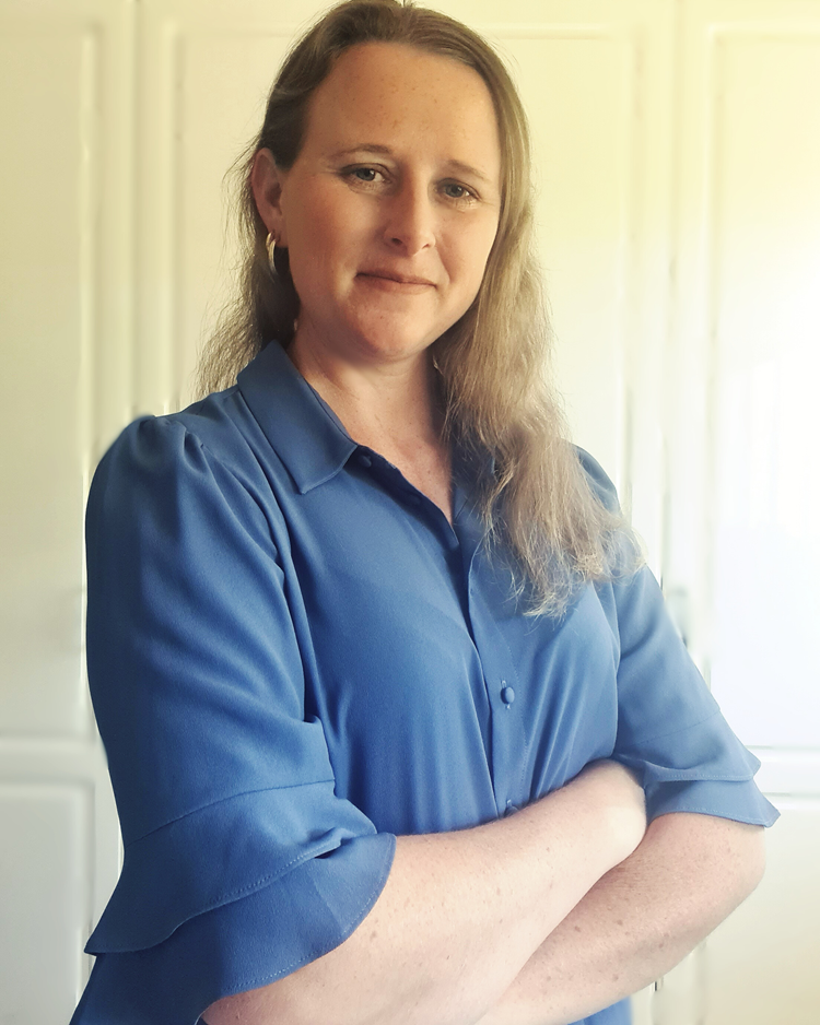 Woman with long blonde hair wearing a blue blouse, standing with arms crossed in a softly lit room.