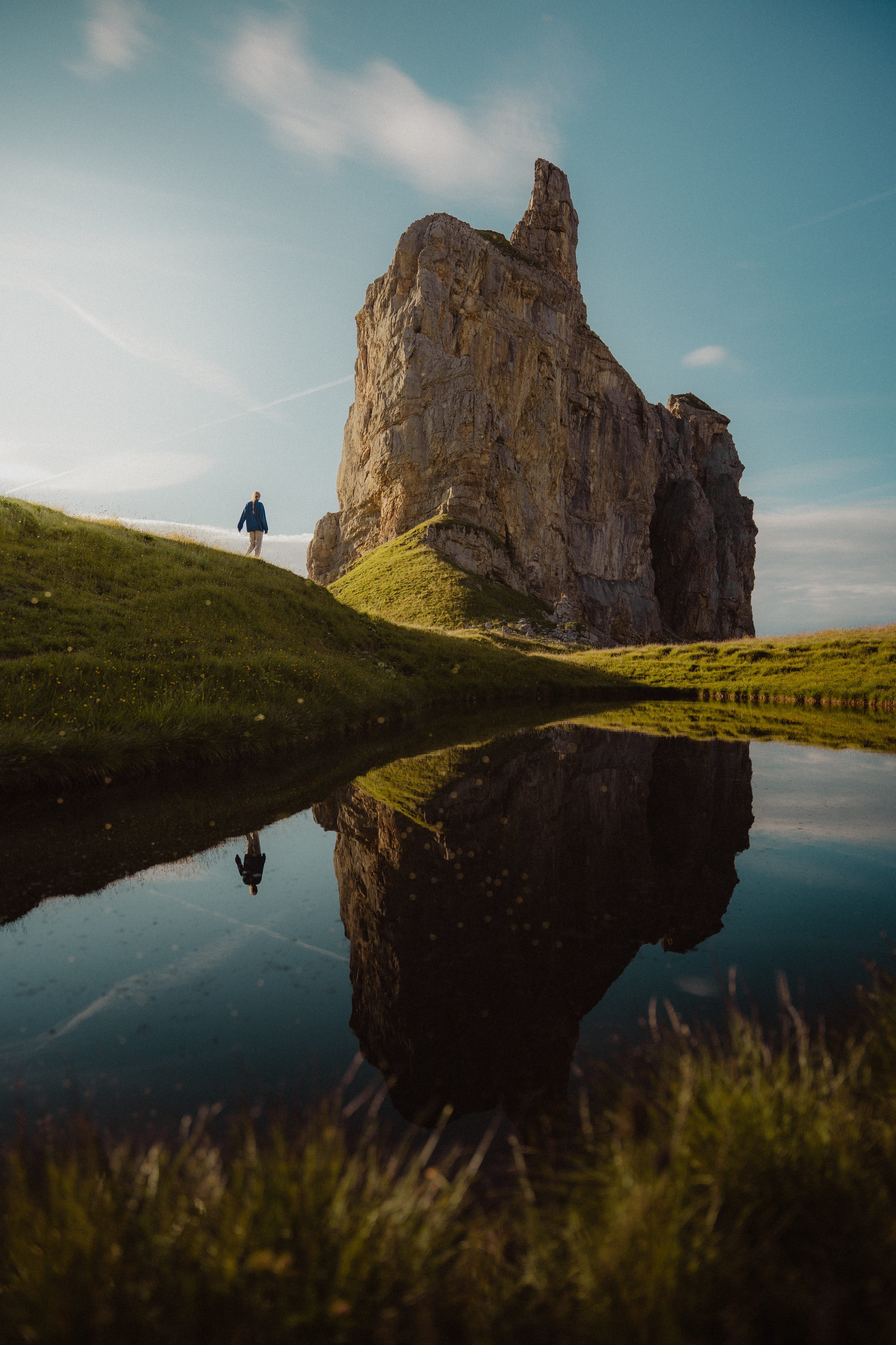 Person standing on a grassy hill beside a large rock formation reflected in a calm pond.