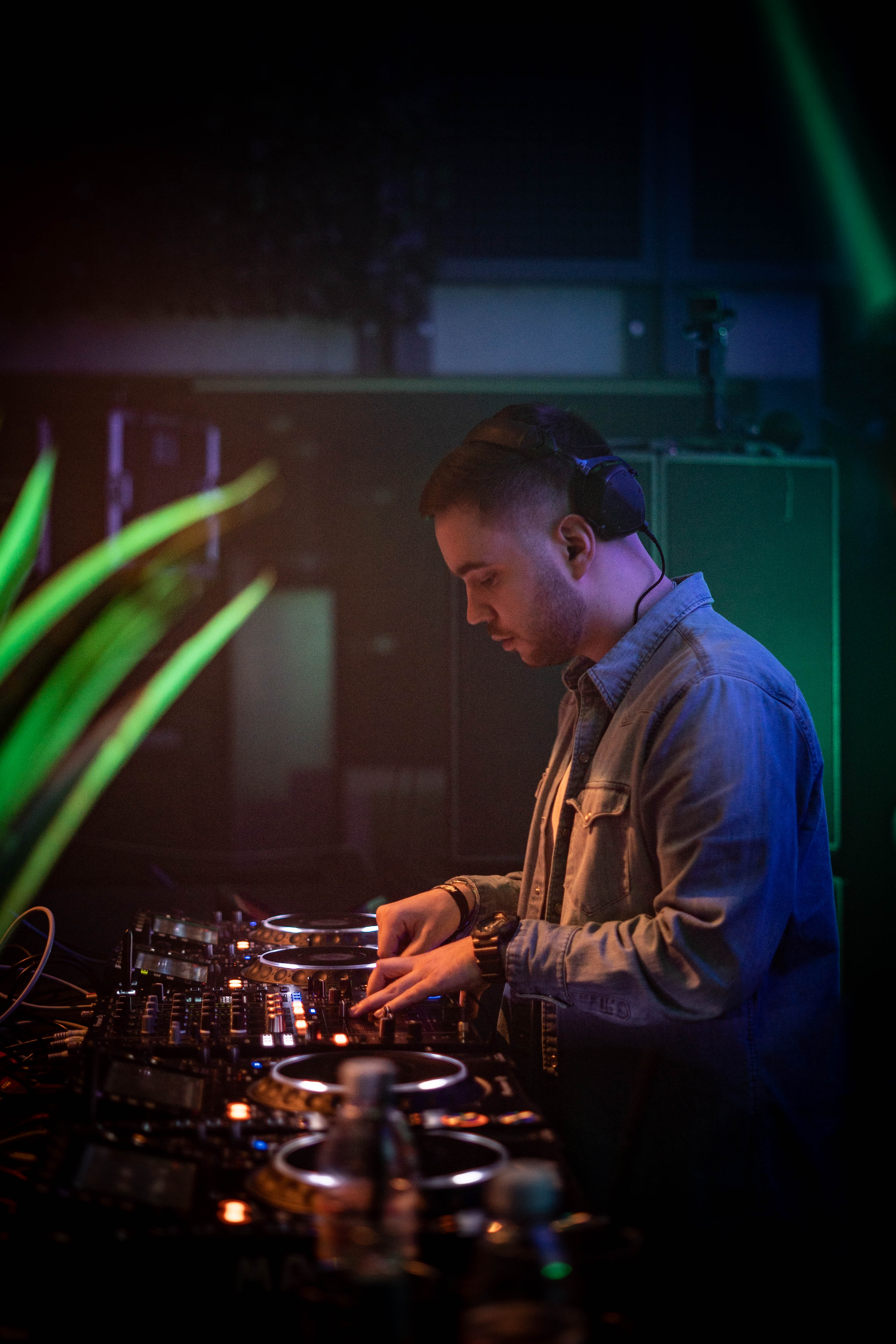 DJ wearing headphones adjusting controls on a mixing console in a dimly lit club or studio.