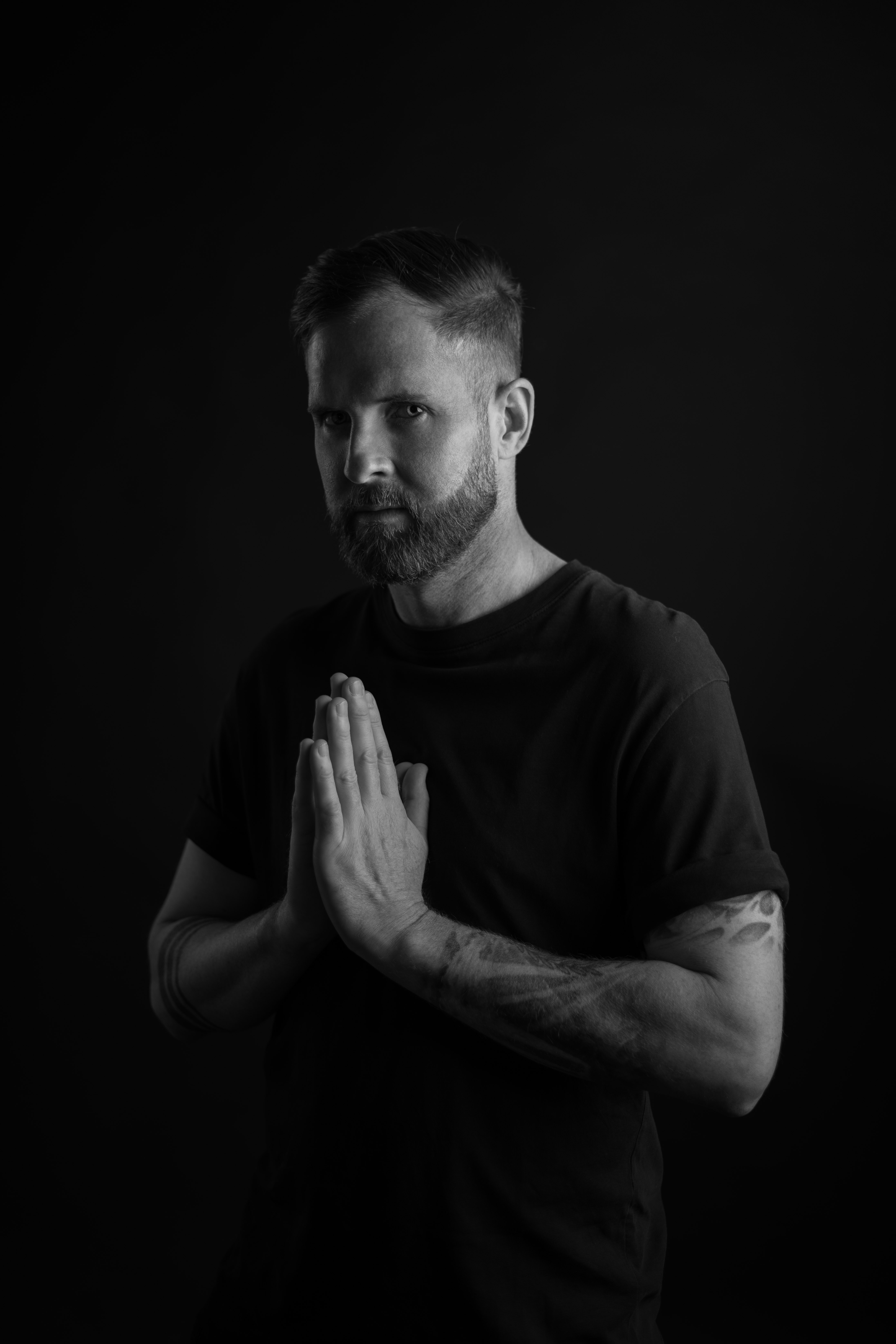 Bearded man with tattooed arms in black t-shirt holds hands pressed together at chest against black background.