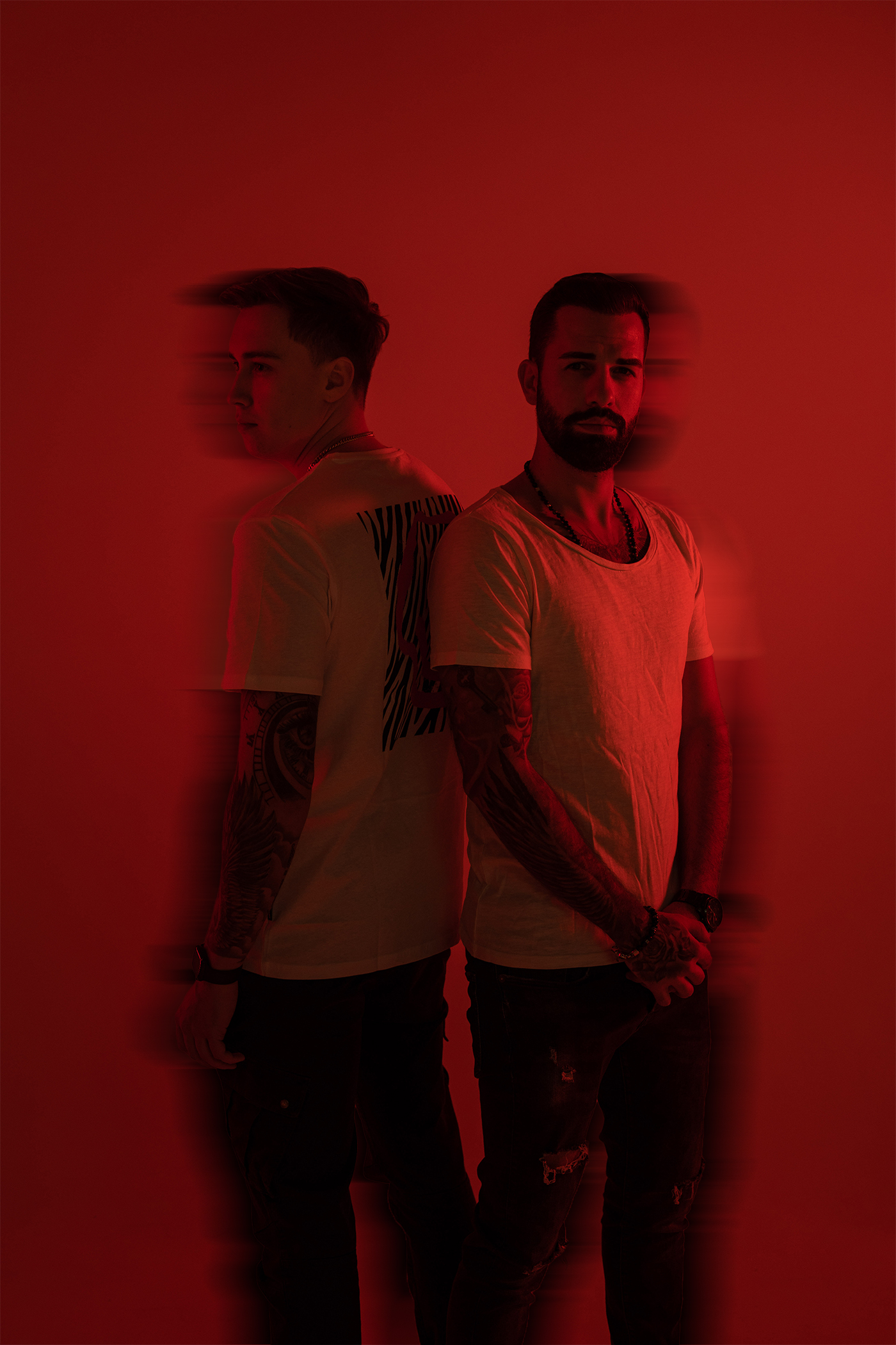 Two tattooed men with beards and short hair stand back to back against a red background with motion blur effect.