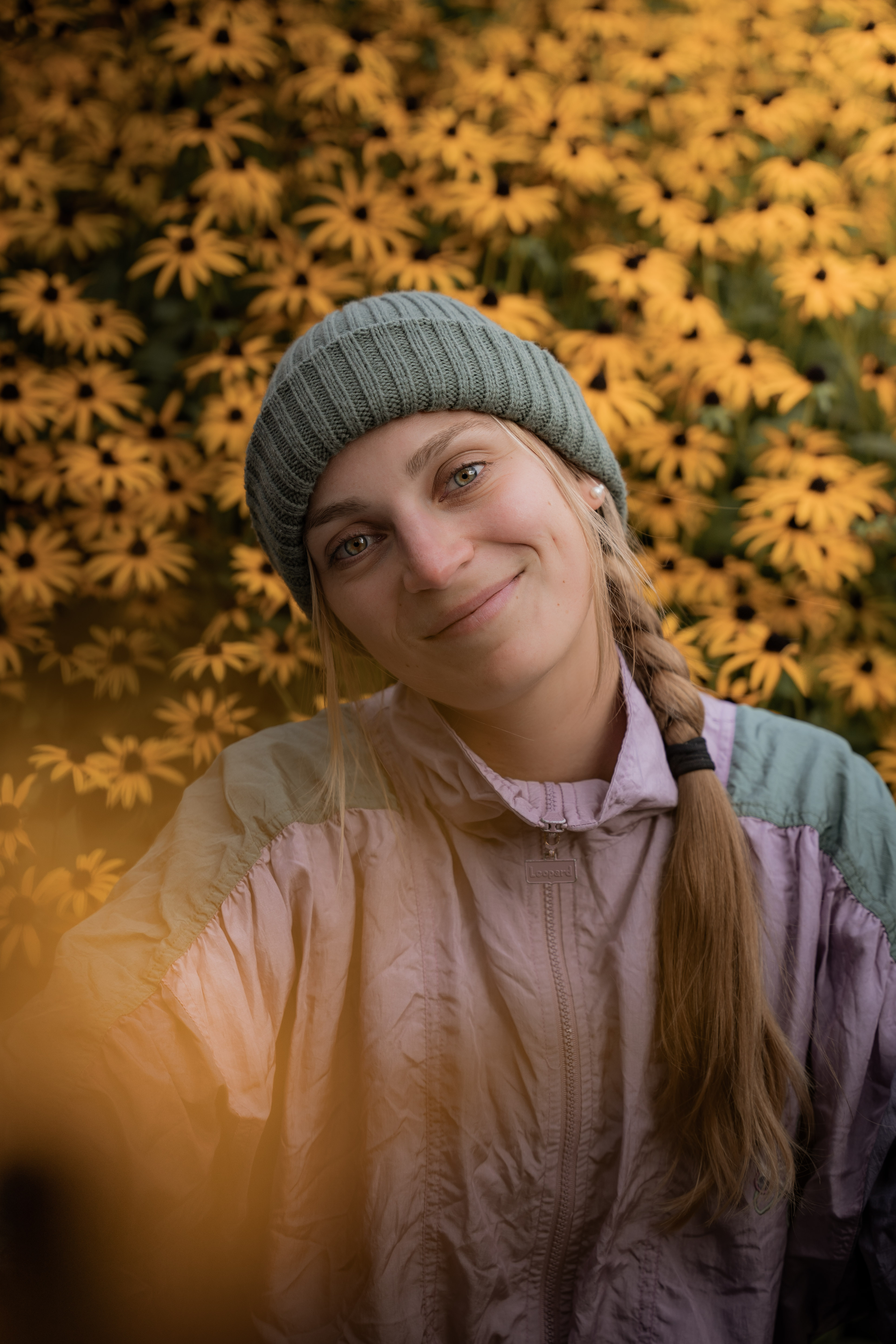 Smiling woman wearing a gray knit beanie and light purple jacket, with long braided hair, standing in front of a backdrop of yellow flowers.