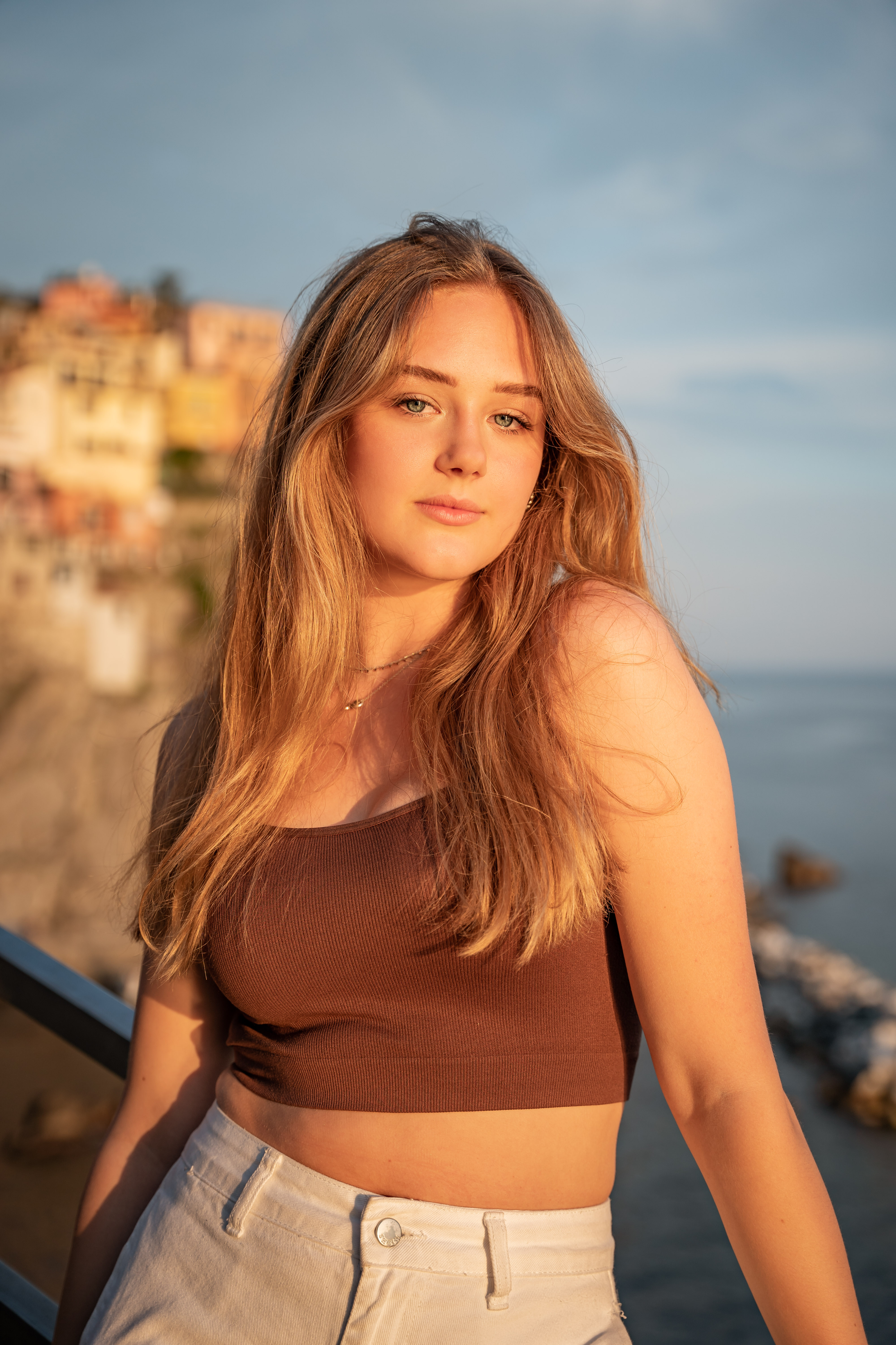 Young woman with long blonde hair wearing a brown crop top and white pants, standing near a seaside cliff during golden hour.