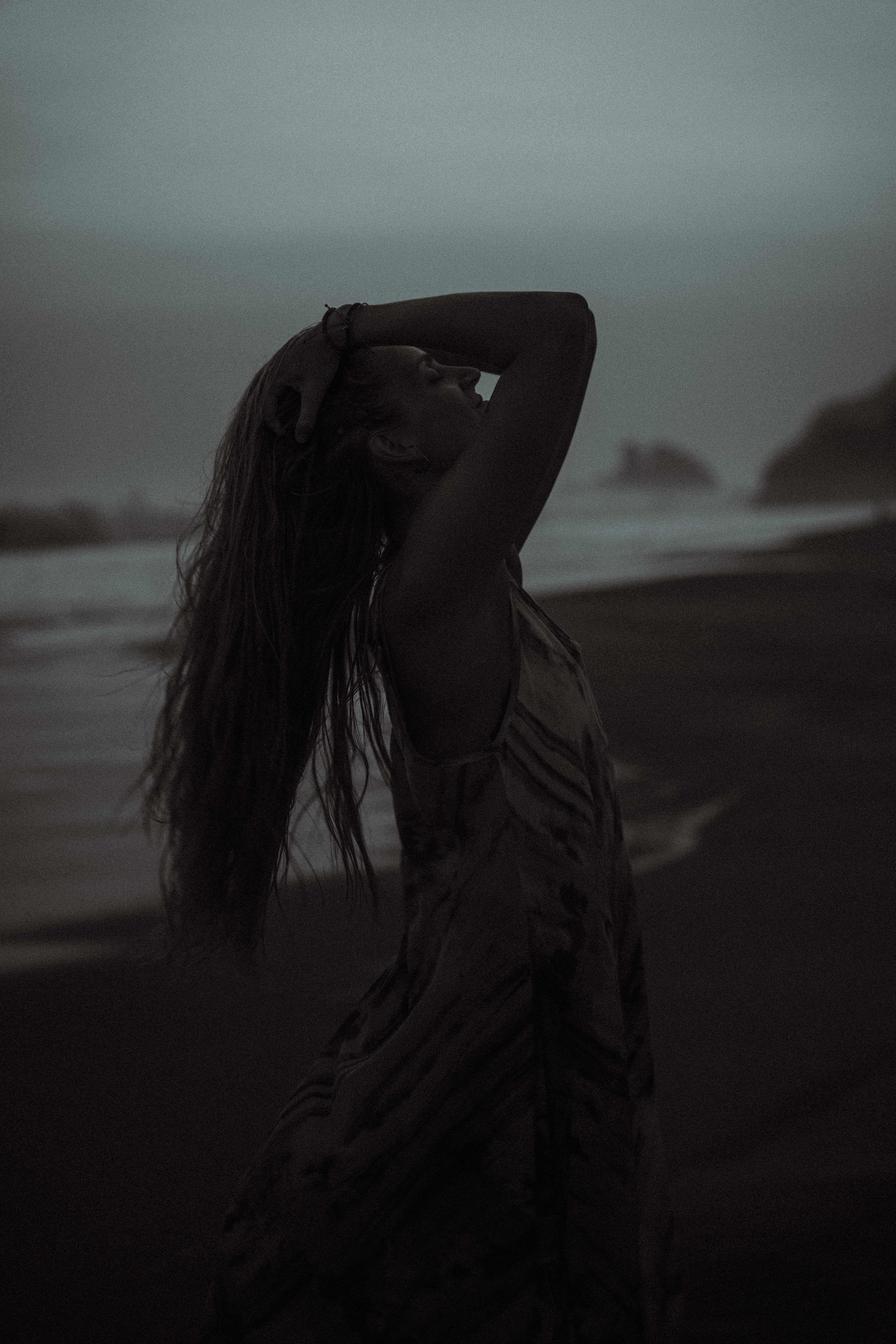 Silhouette of a woman with long hair holding her head on a beach at dusk.
