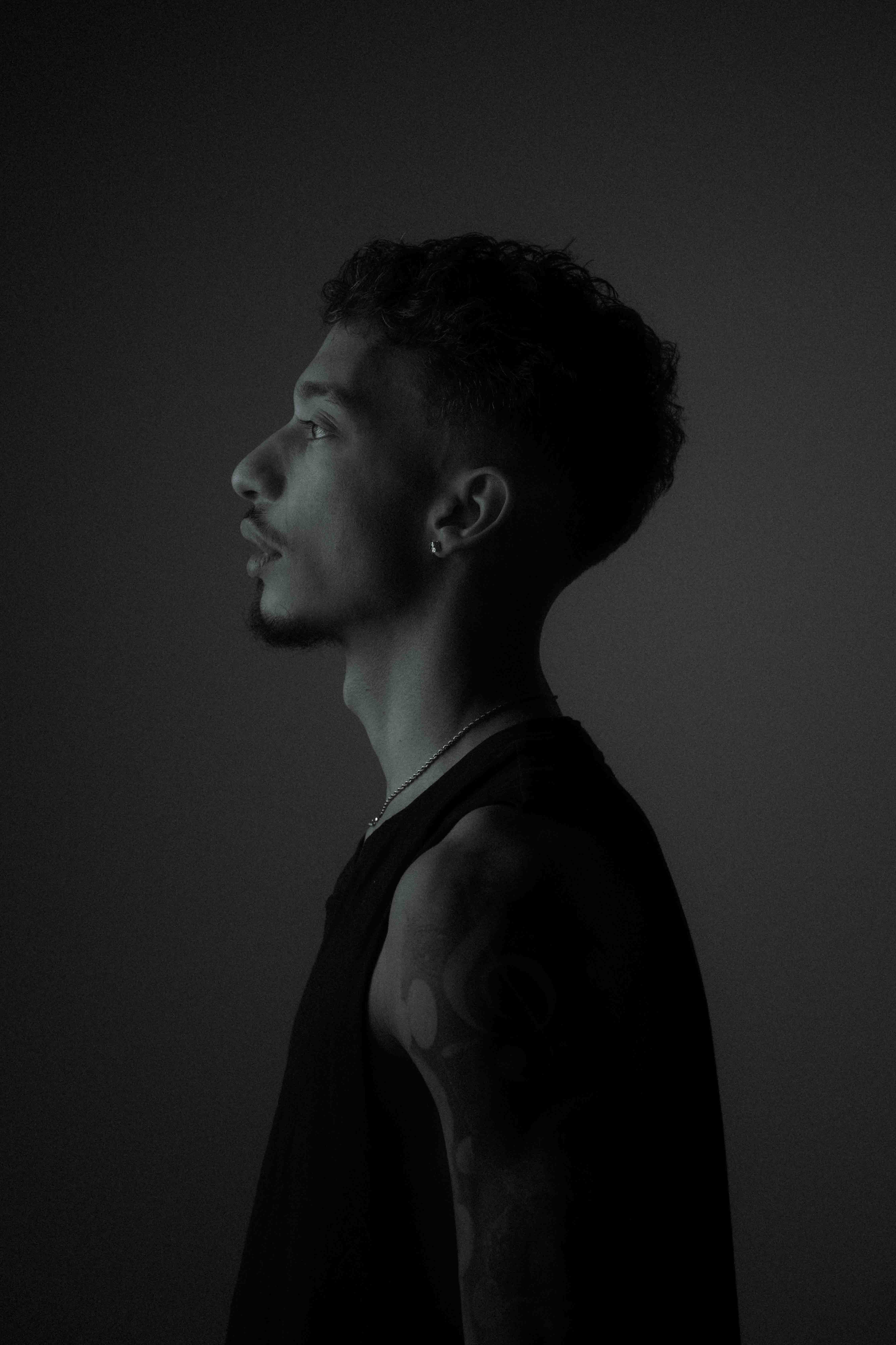 Black and white profile portrait of a young man with curly hair, an earring, and a tattoo on his arm against a dark background.