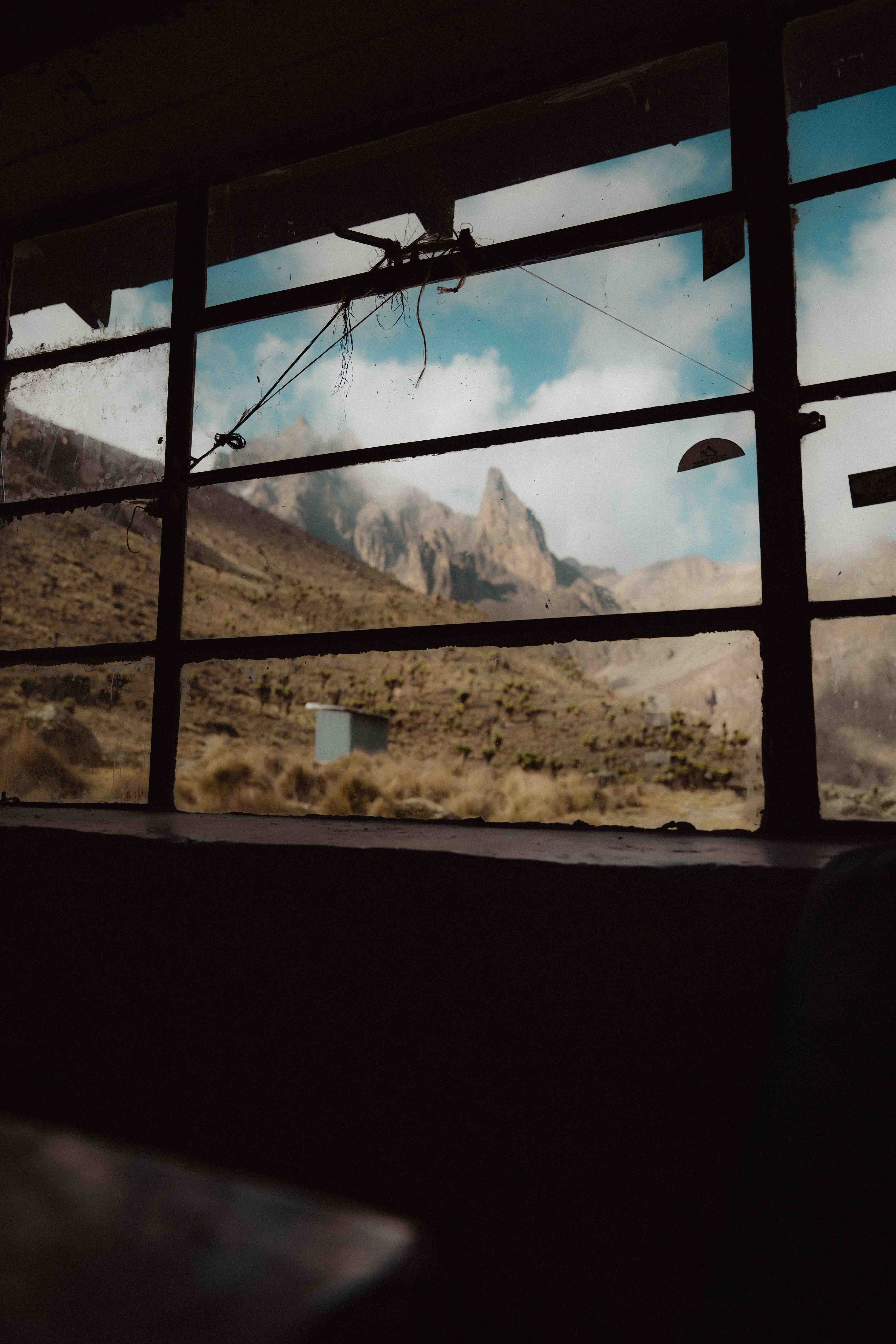 View through a dirty window showing a dry mountainous landscape with a small building and a partly cloudy sky.