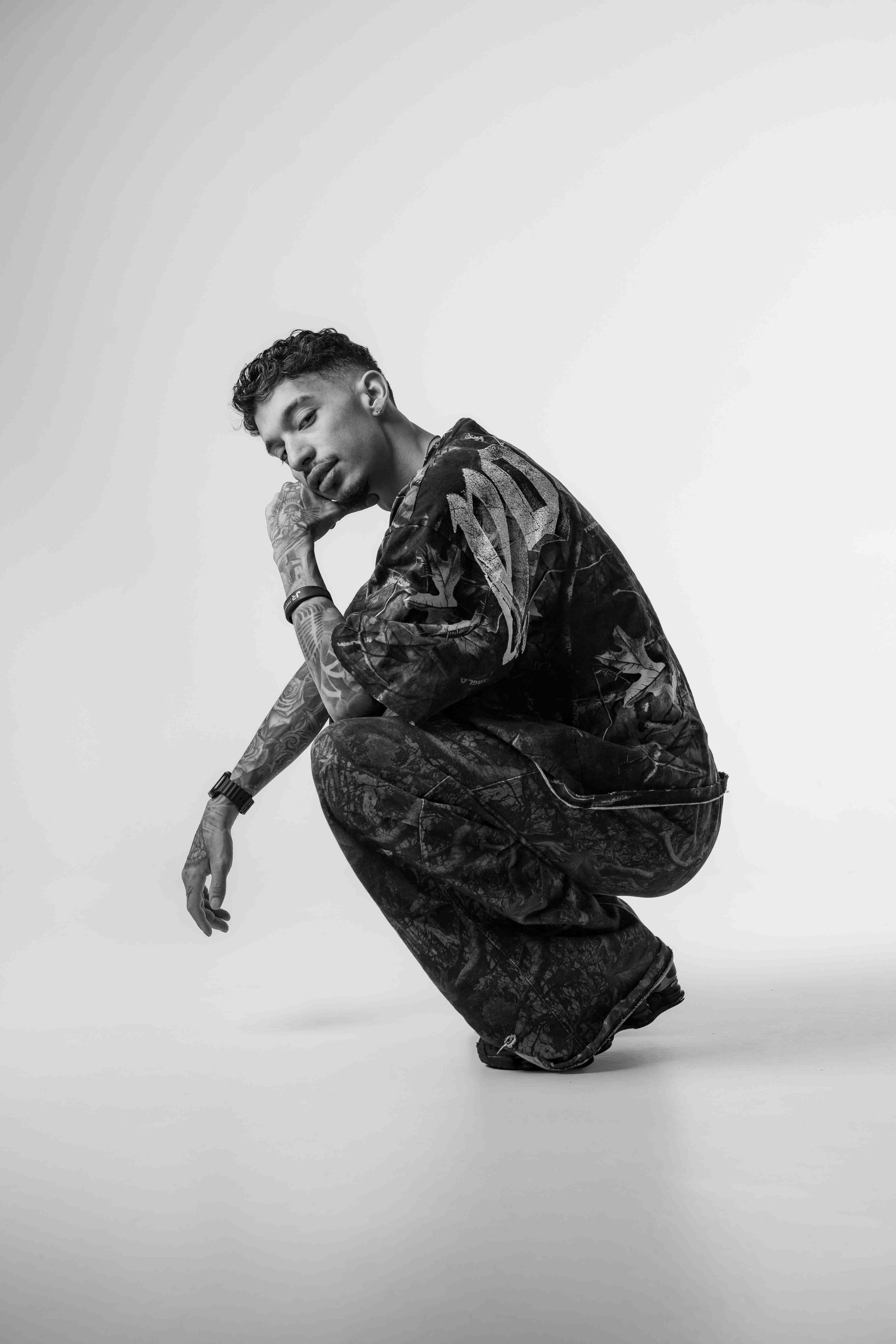 Young man with tattoos squatting, resting his chin on his hand, against a plain white background in black and white.