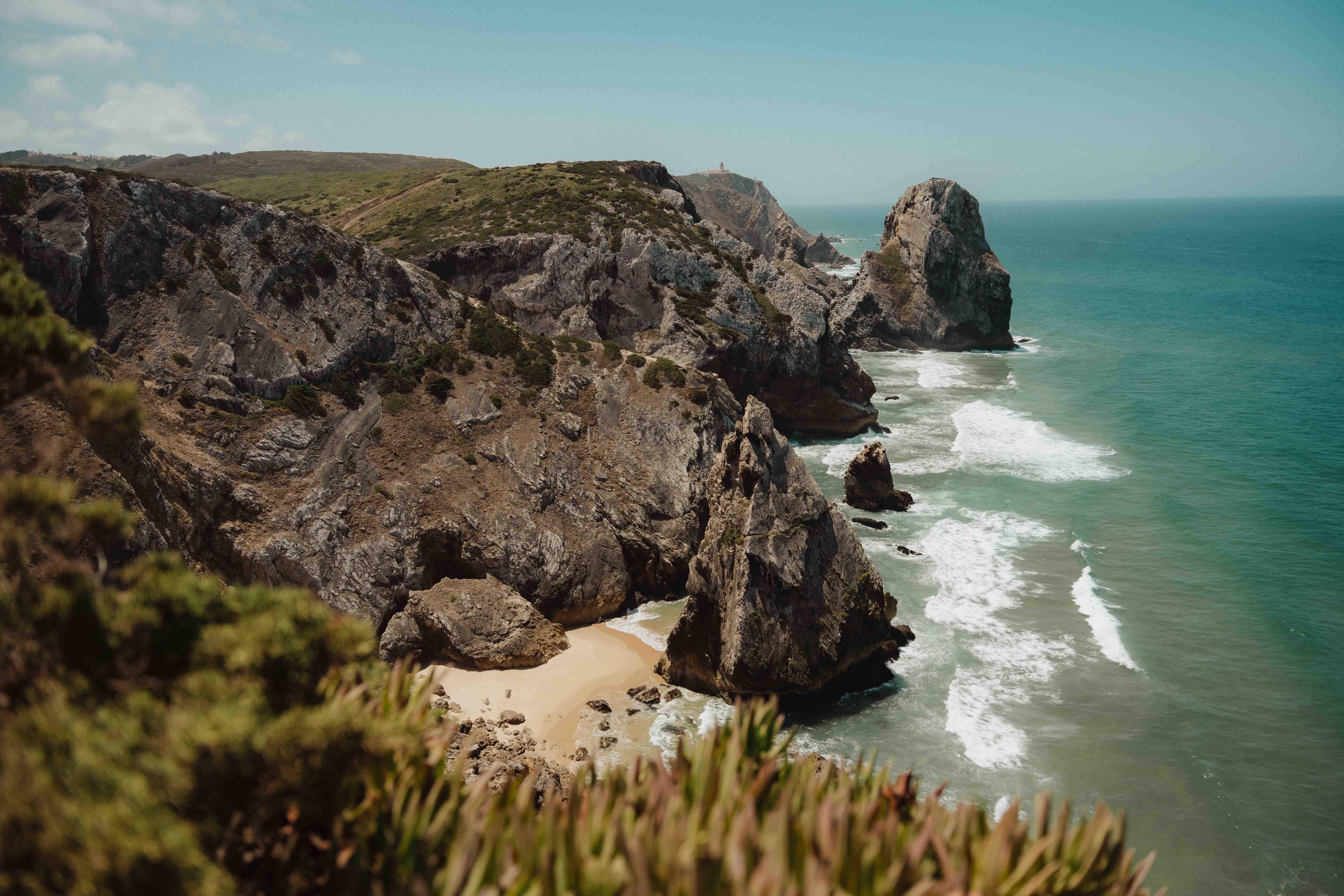 Rocky cliffs and large rock formations along a coastline with ocean waves and a small sandy beach.