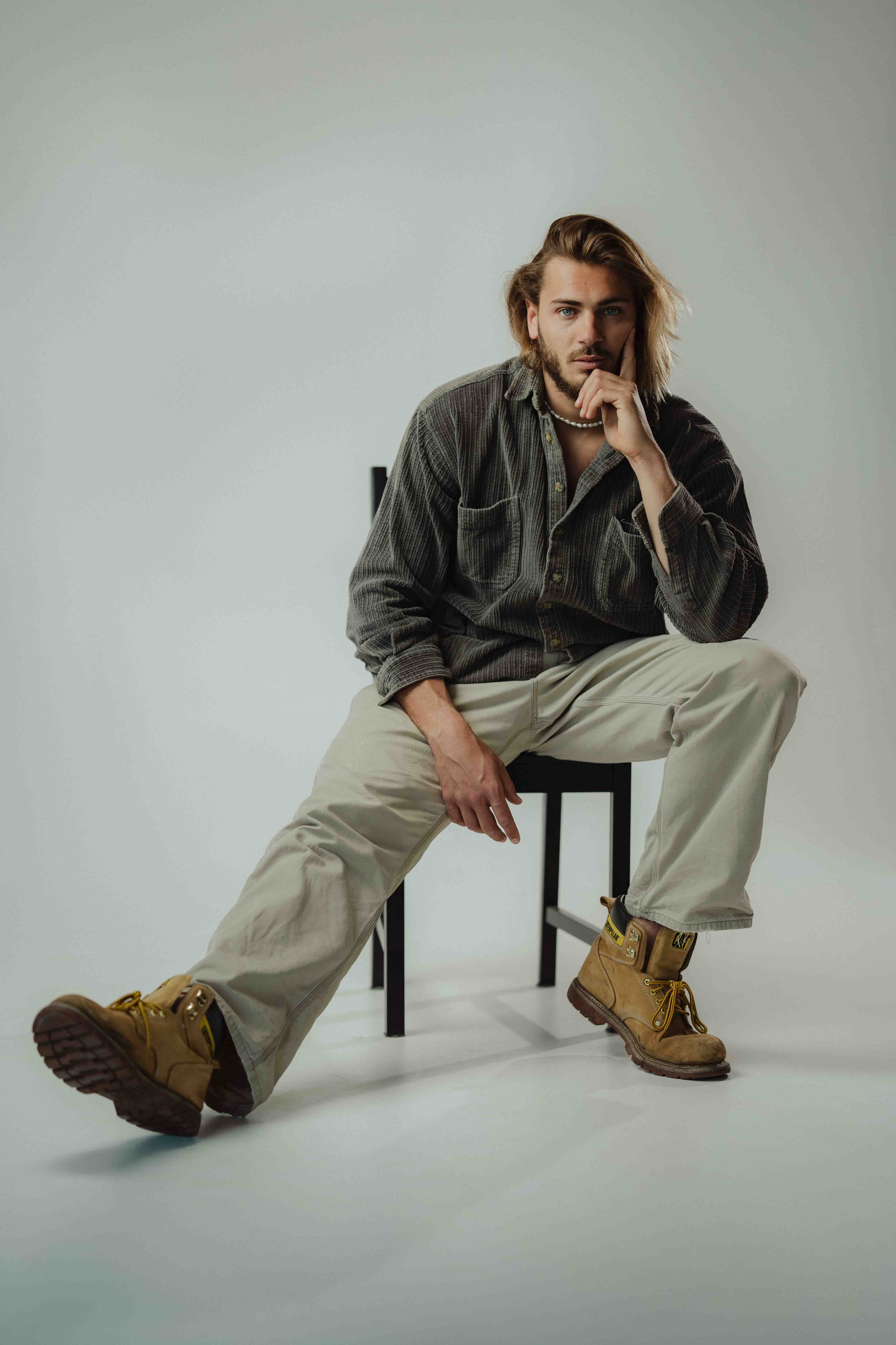 Man with long hair wearing a dark shirt, beige pants, and tan work boots sitting on a black chair with one leg stretched out.
