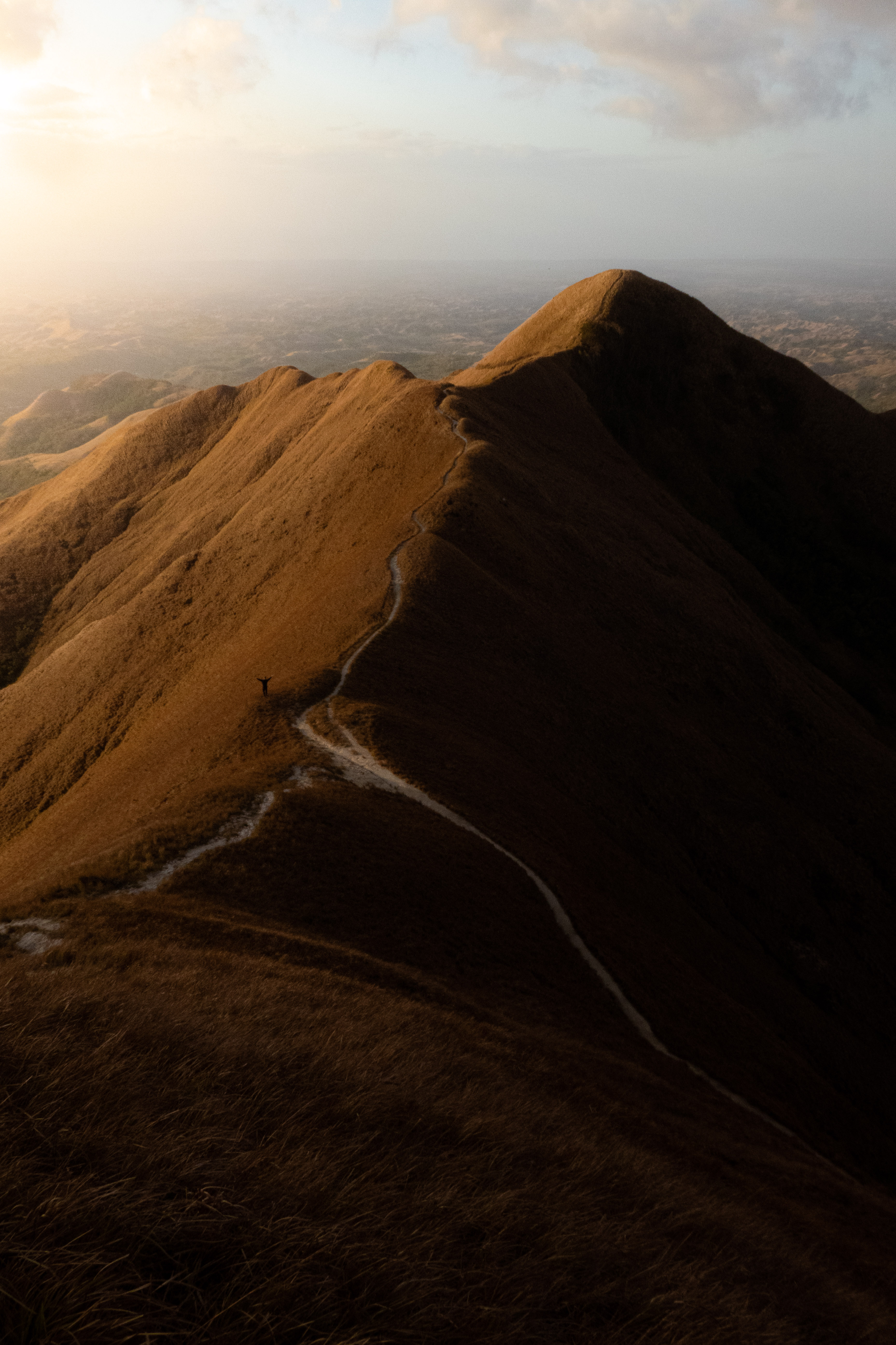 Trail winding up a sunlit mountain ridge with a person standing and raising arms.