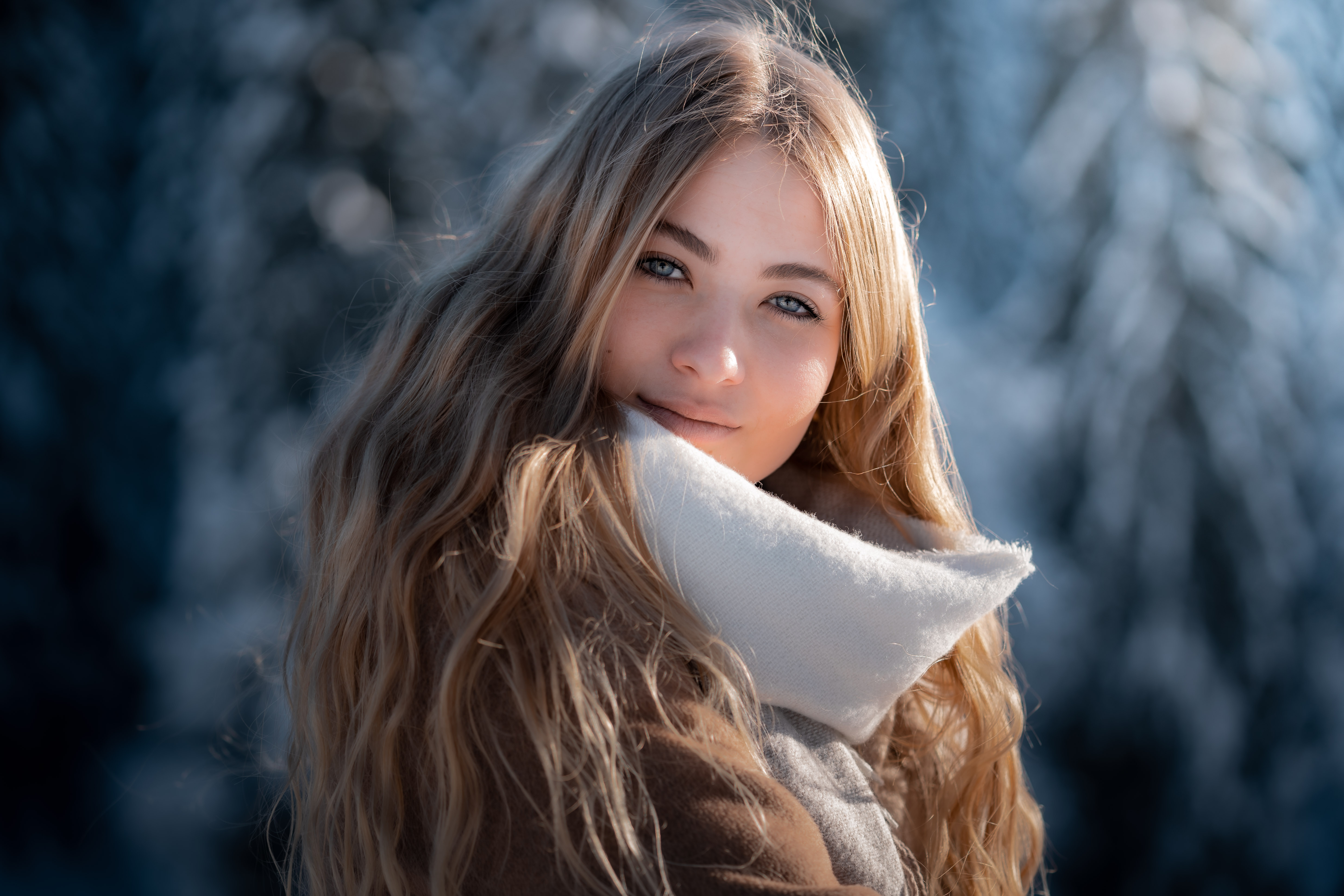 Young woman with long wavy blonde hair wearing a warm coat and white scarf in a snowy outdoor setting.