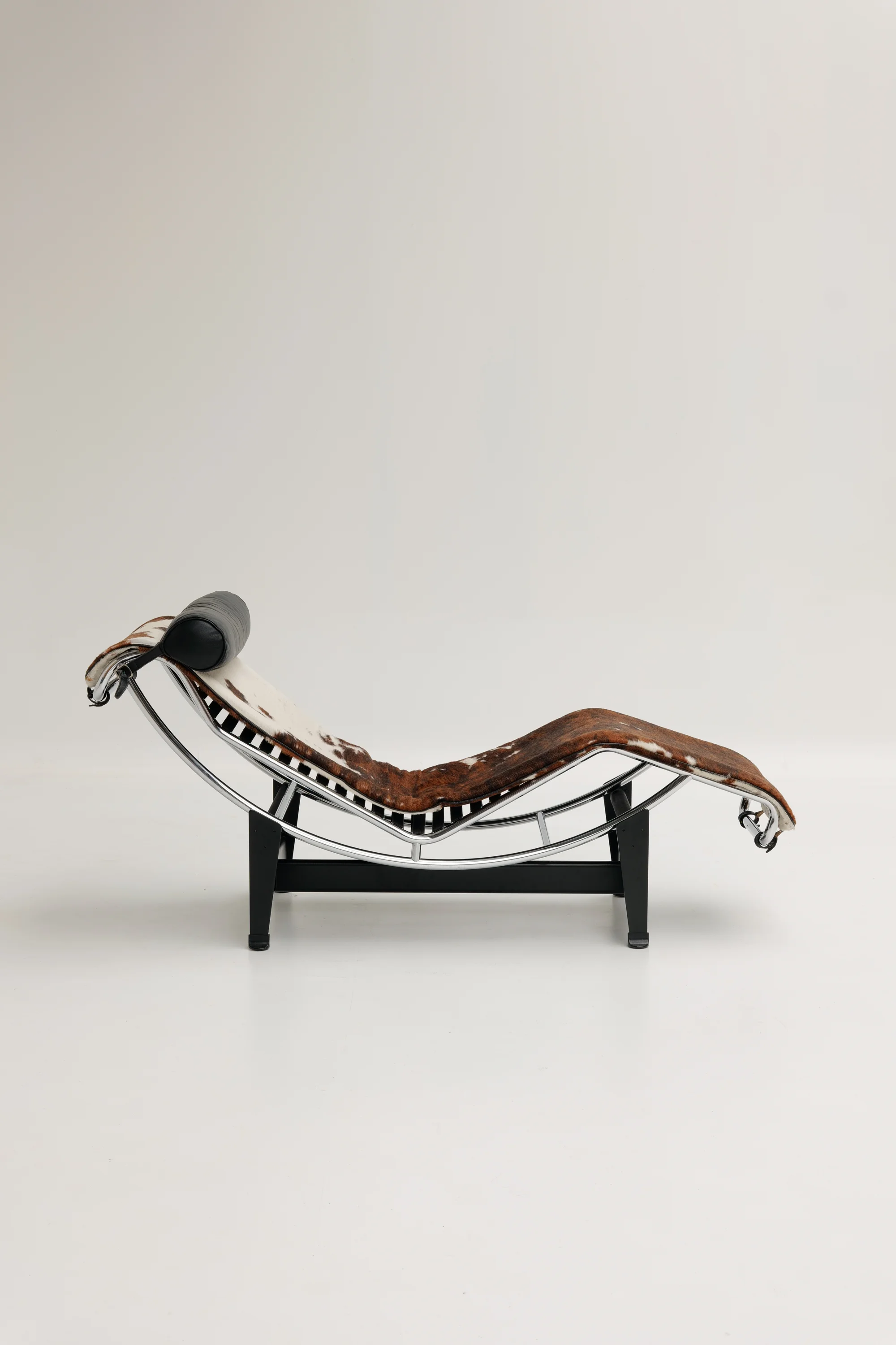 Modern chaise lounge chair with a curved metal frame and cowhide upholstery, featuring a black headrest pillow.