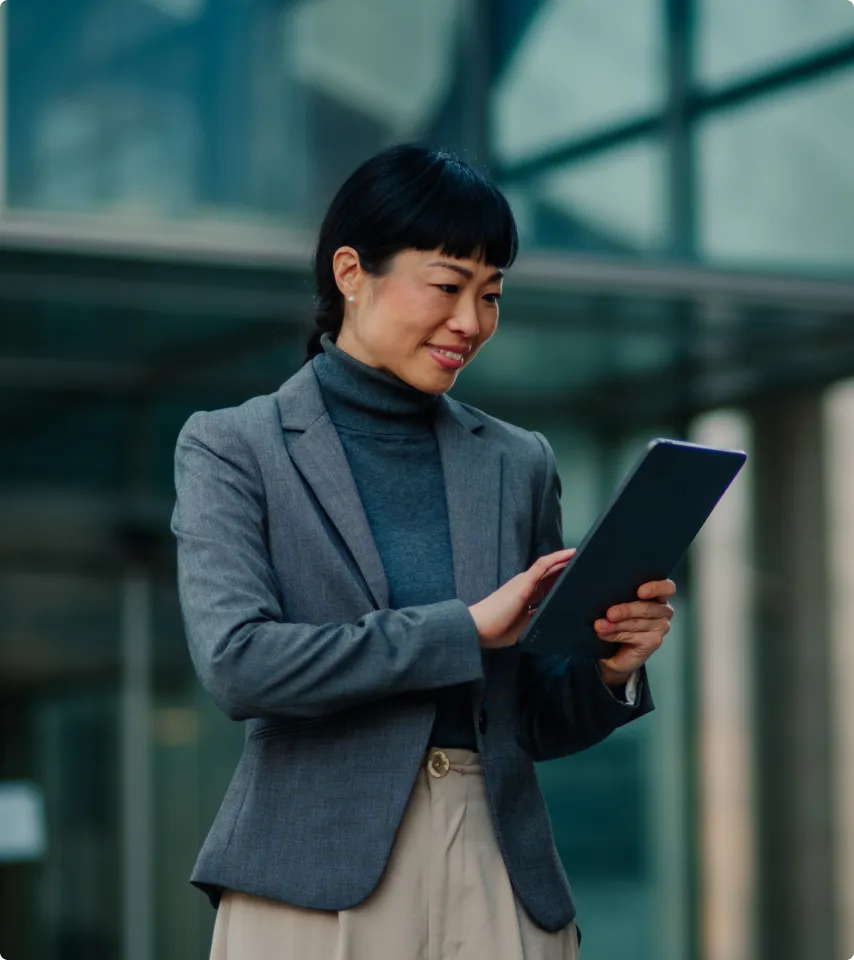 Smiling woman in a gray blazer using a digital tablet outdoors in front of a modern building.