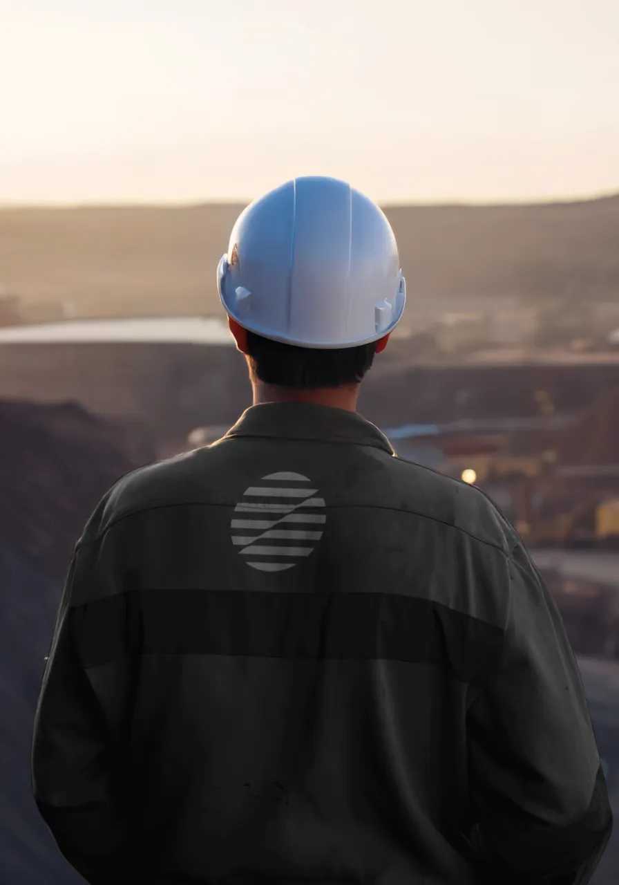 Person wearing a white safety helmet and dark jacket looking out over an open-pit mining site at sunset.