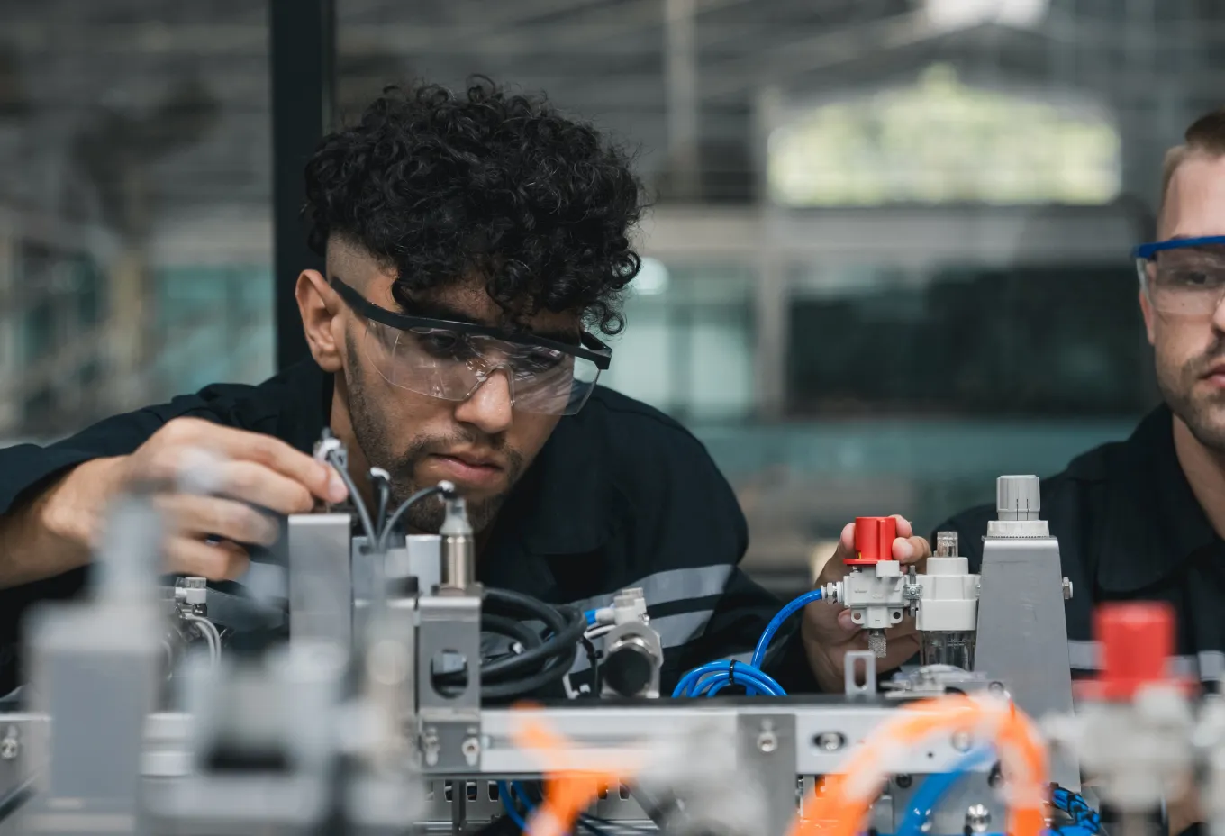 Two male technicians wearing safety glasses working on complex industrial machinery with pipes and wires.