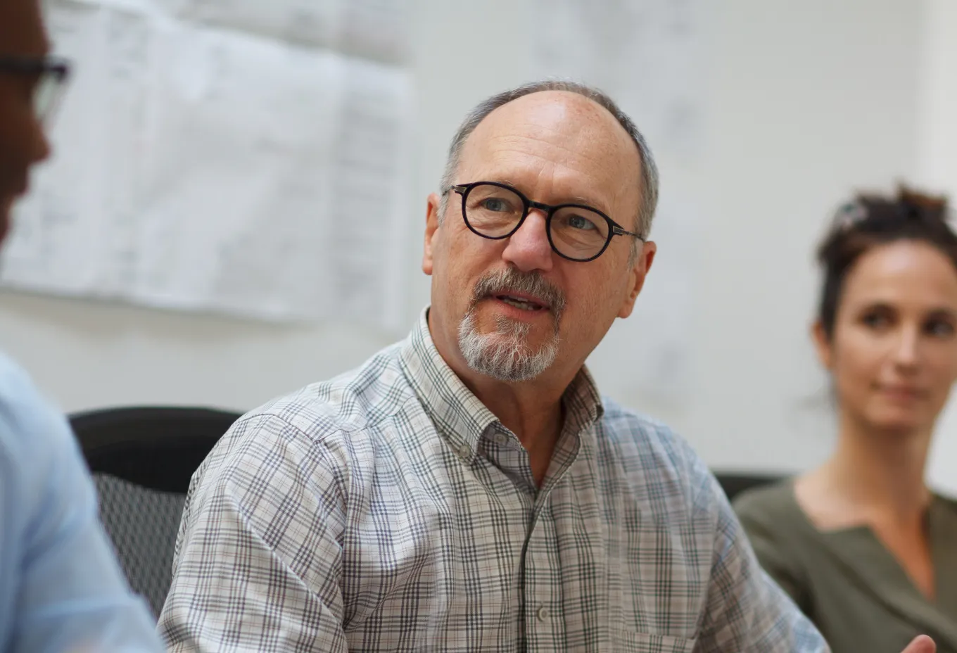 Middle-aged man with glasses and a goatee speaking in a meeting with two blurred colleagues in the background.