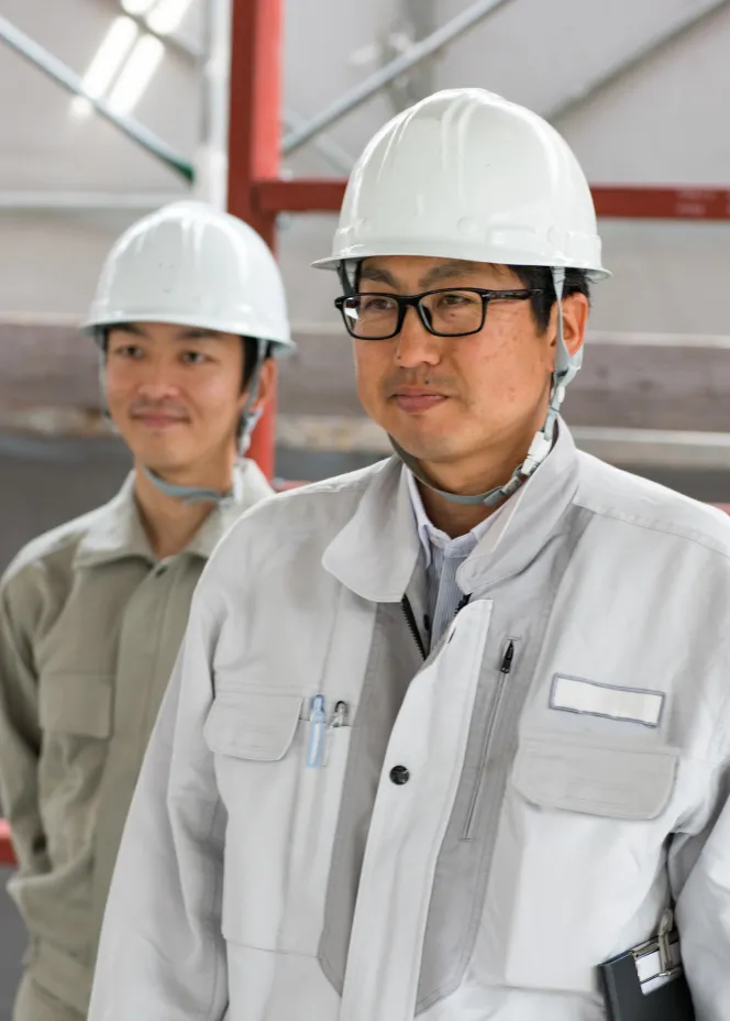 Two male construction workers wearing white hard hats and work jackets in an indoor industrial setting.