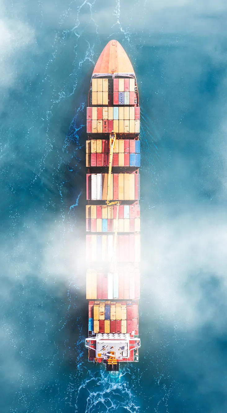 Aerial view of a large cargo ship loaded with colorful shipping containers navigating through blue ocean waters with mist partially obscuring the center.