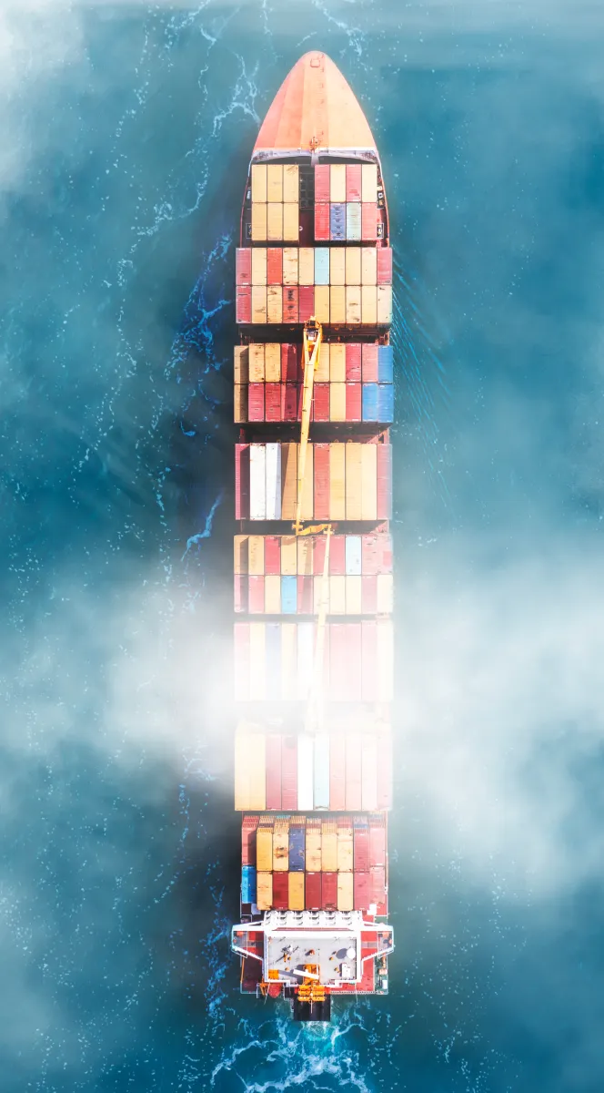 Aerial view of a large cargo ship loaded with colorful shipping containers navigating through blue ocean waters with mist partially obscuring the center.