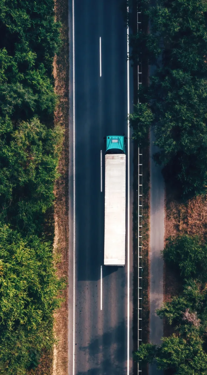 Aerial view of a teal semi-truck with a white trailer driving on a two-lane highway surrounded by green trees.