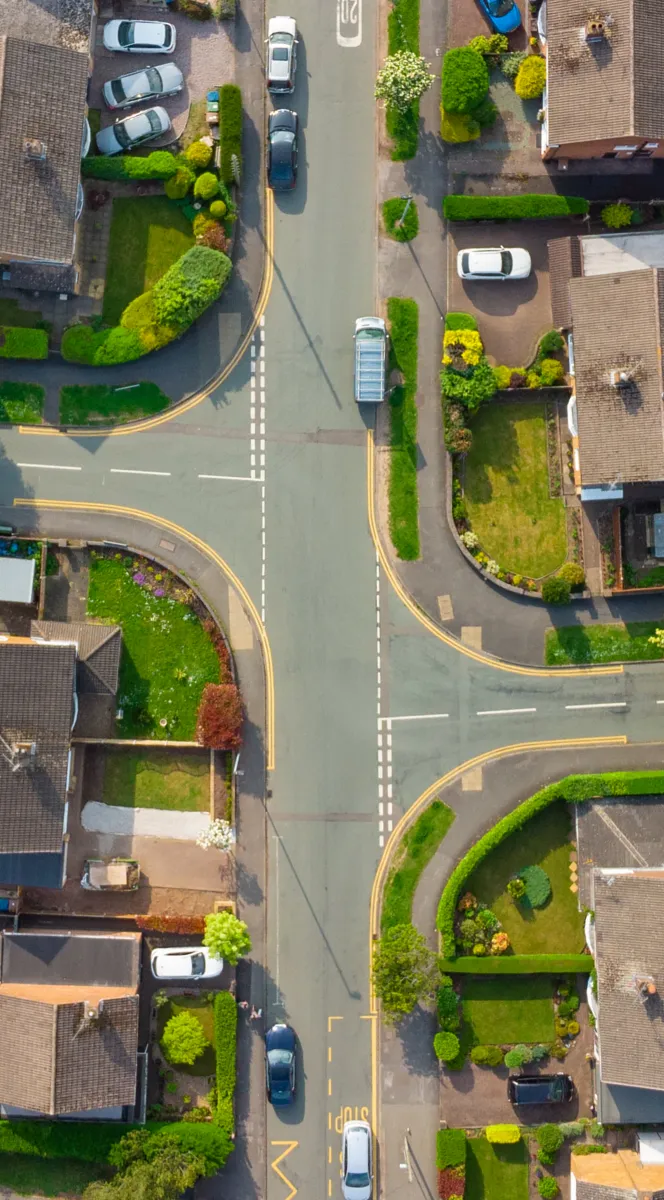 Aerial view of a residential intersection with cars, houses, green lawns, and well-maintained gardens.