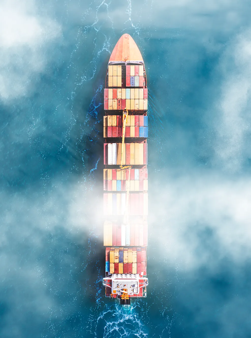 Aerial view of a large cargo ship with colorful shipping containers sailing through blue water partly covered by mist.