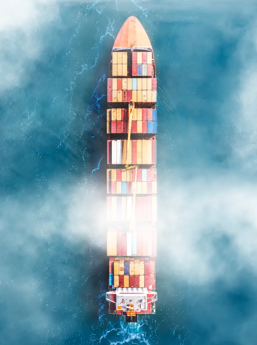 Aerial view of a large cargo ship with colorful shipping containers sailing through blue water partly covered by mist.