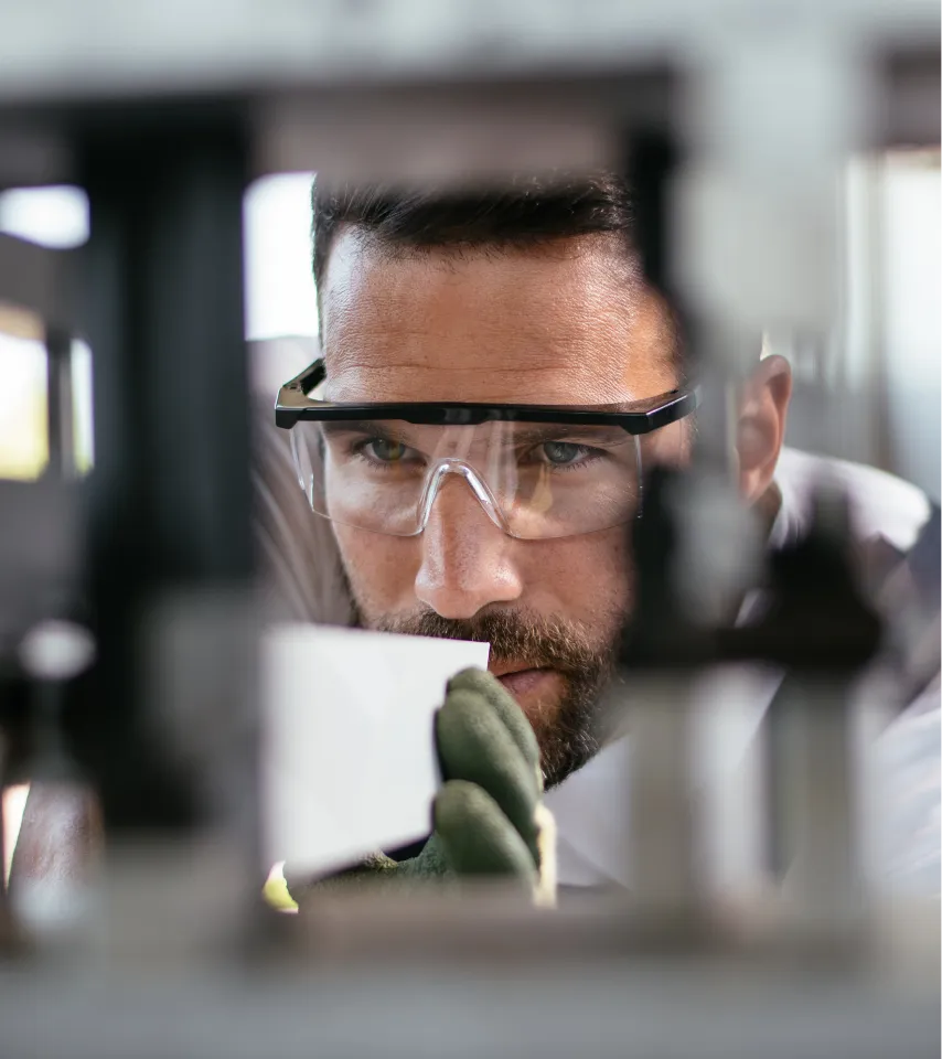 Man wearing safety glasses and gloves closely inspecting a small white object in a workshop.