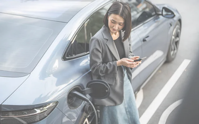 Woman in blazer standing next to an electric car that is charging, looking at her phone.