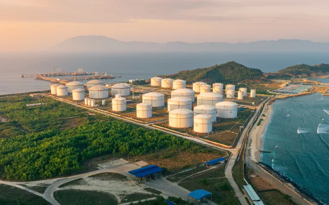 Aerial view of a coastal oil storage facility with multiple large white cylindrical tanks near green vegetation and the ocean at sunset.
