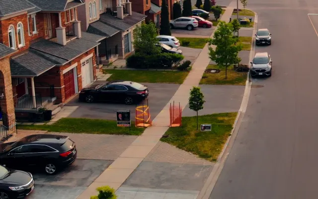 Suburban street with parked cars, brick townhouses, small trees, and political campaign signs along the sidewalk.